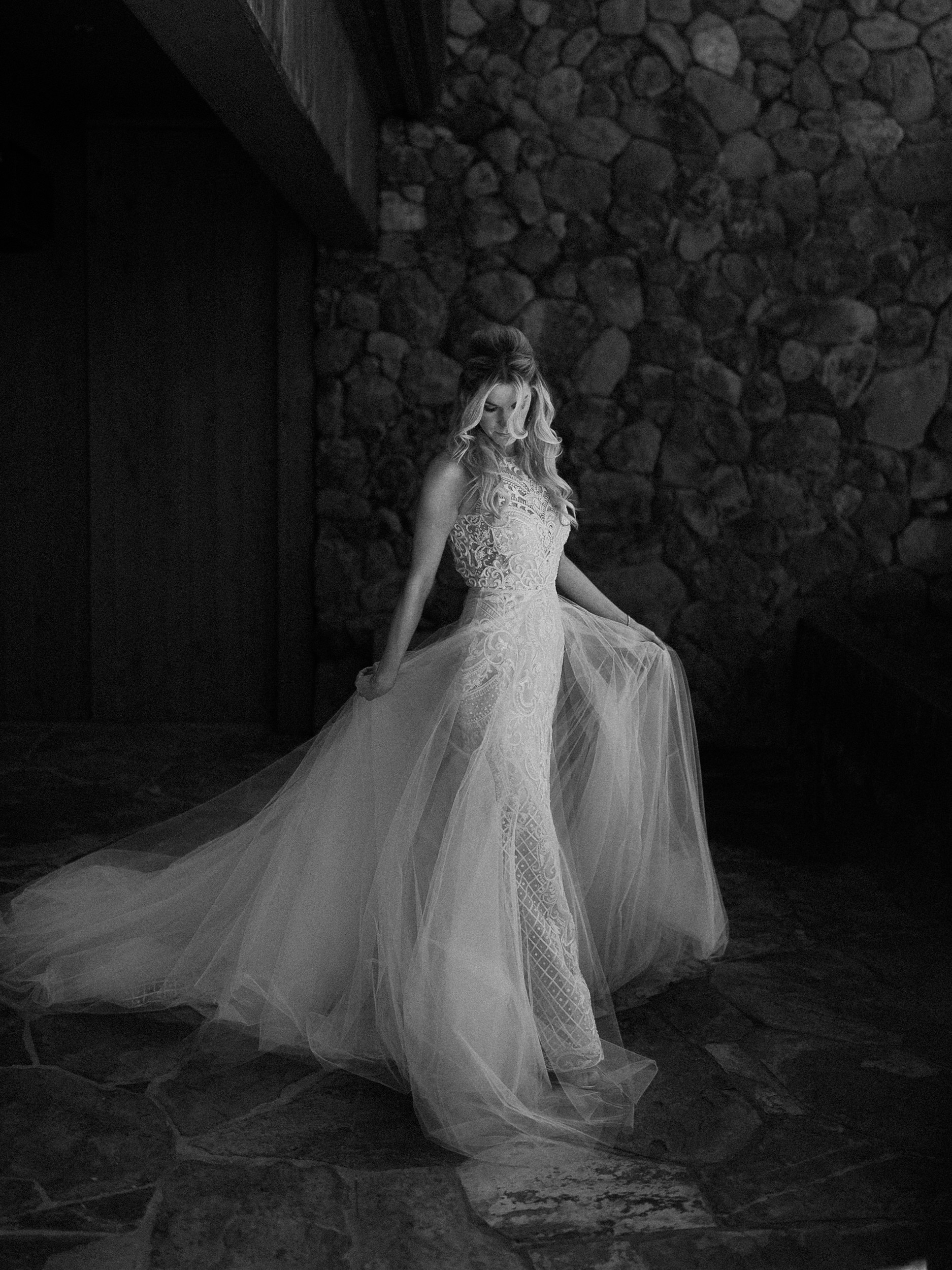 Bride in lace dress stands indoors on a stone floor, holding her gown and looking down, with a stone wall behind. Black and white.