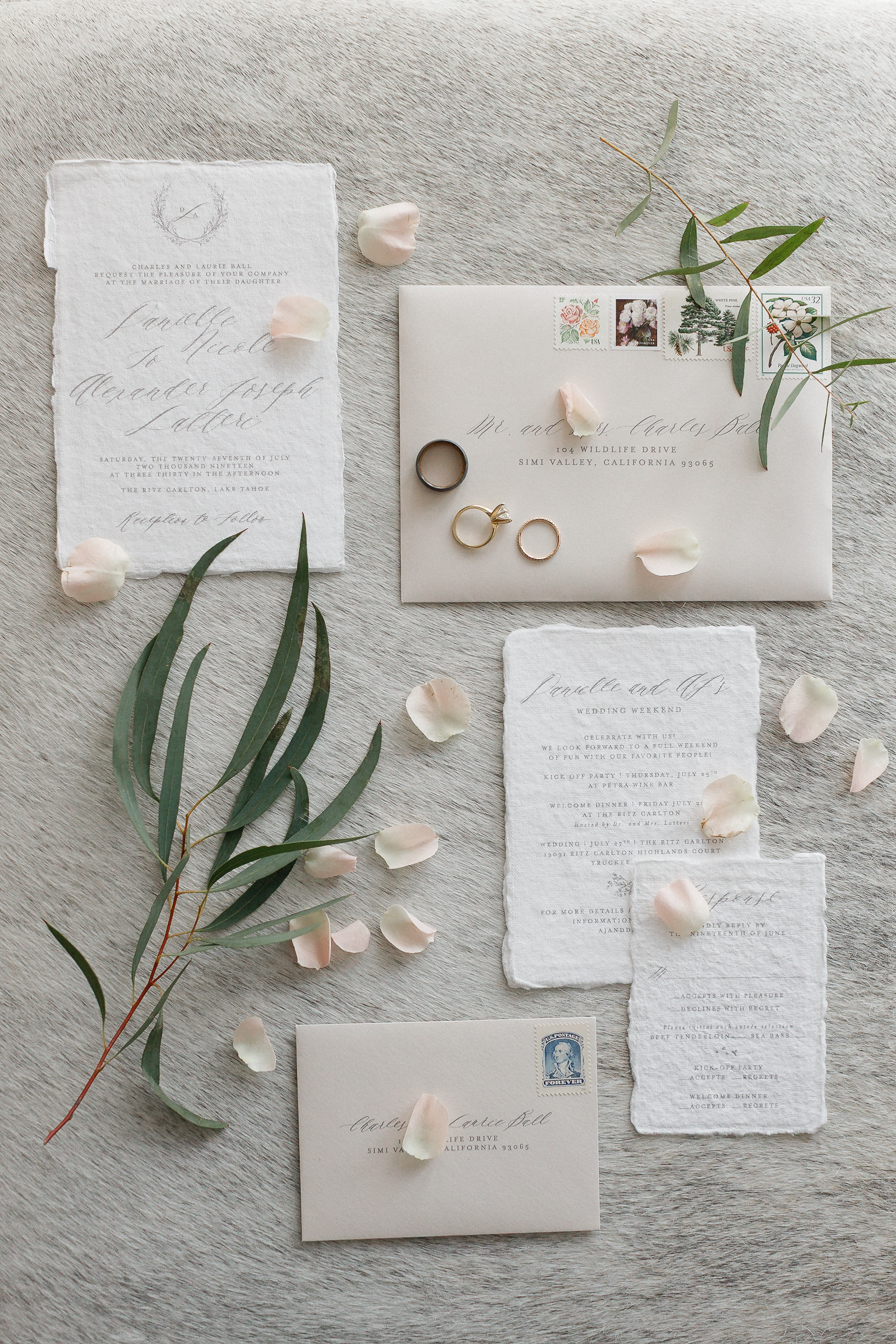 Flat lay of Ritz-Carlton wedding invitations, two envelopes, three rings, rose petals, and eucalyptus on a textured gray surface.