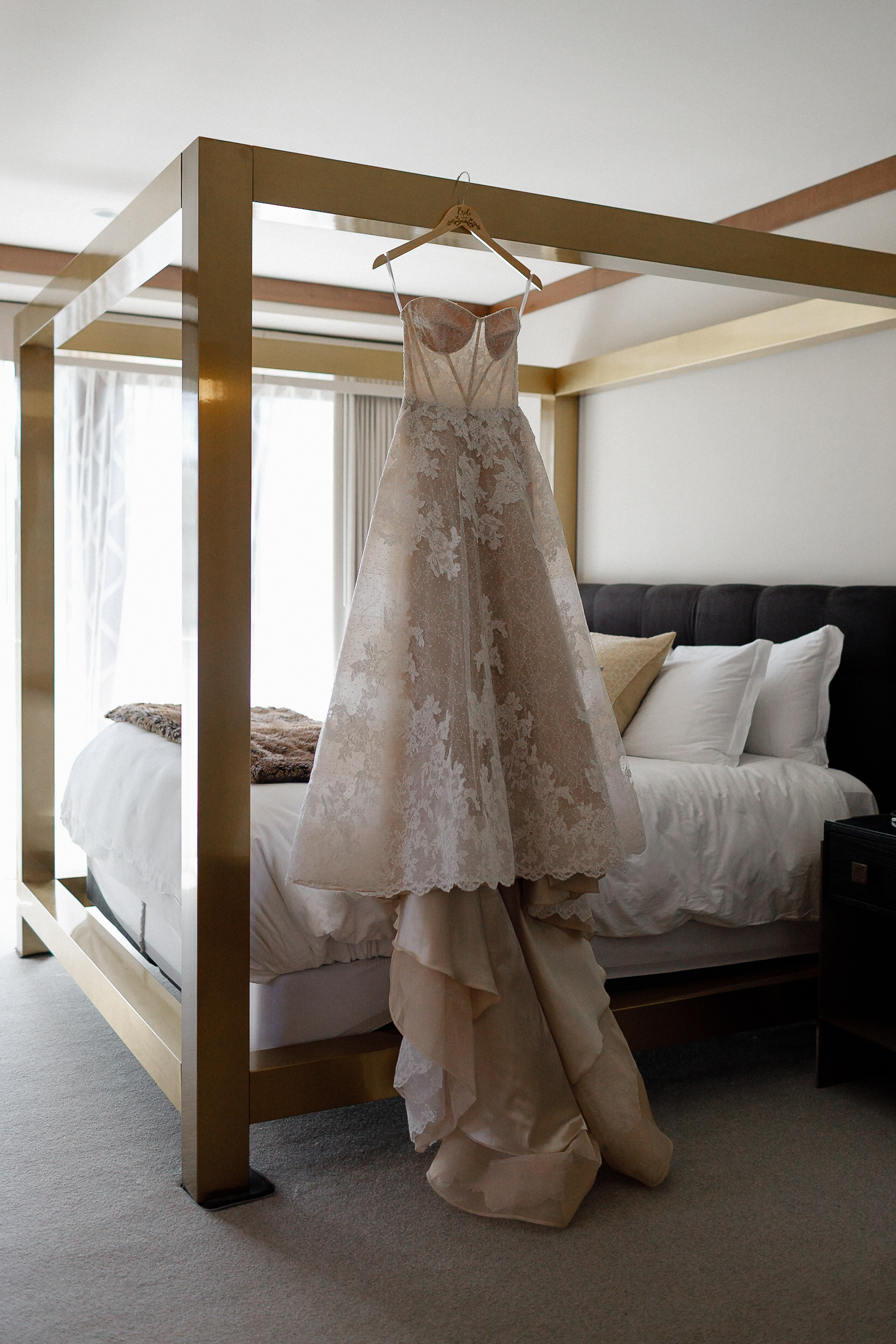 Strapless lace wedding dress with floral pattern on gold four-poster bed in bright Ritz-Carlton room, sunlight on train.