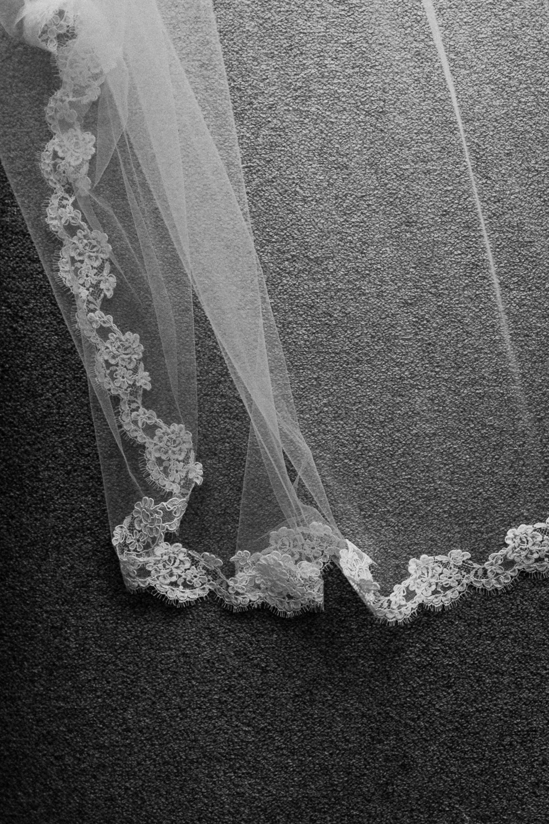 Close-up of a sheer bridal veil with detailed lace trim, laid out on a textured carpeted floor.