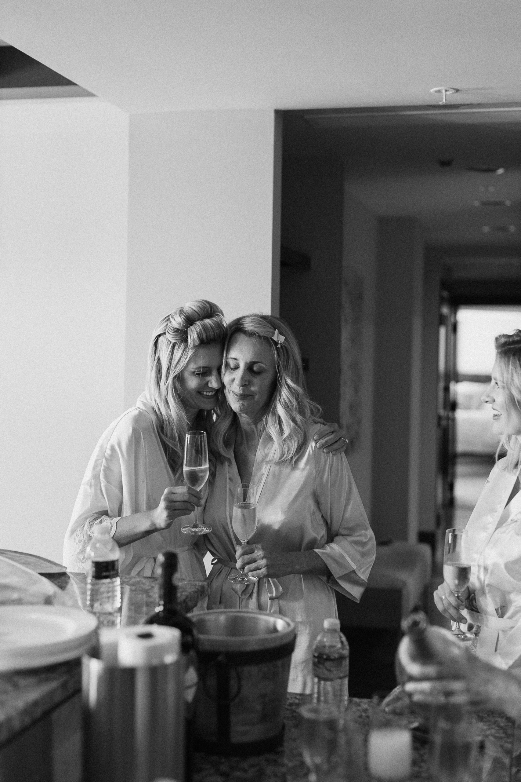 Two women in robes hug and smile with champagne glasses in a softly lit room; a third woman stands nearby, also holding a glass.