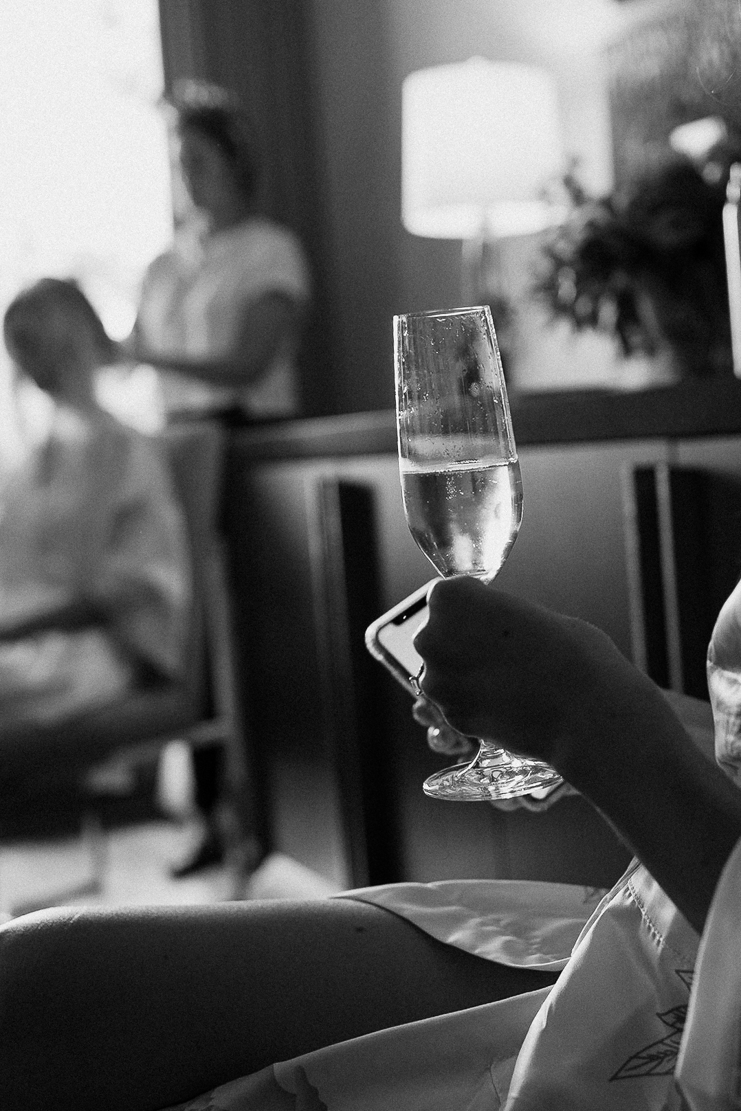 Someone in a robe at the Ritz-Carlton Lake Tahoe holds a champagne flute and phone as another styles hair in a softly lit room.
