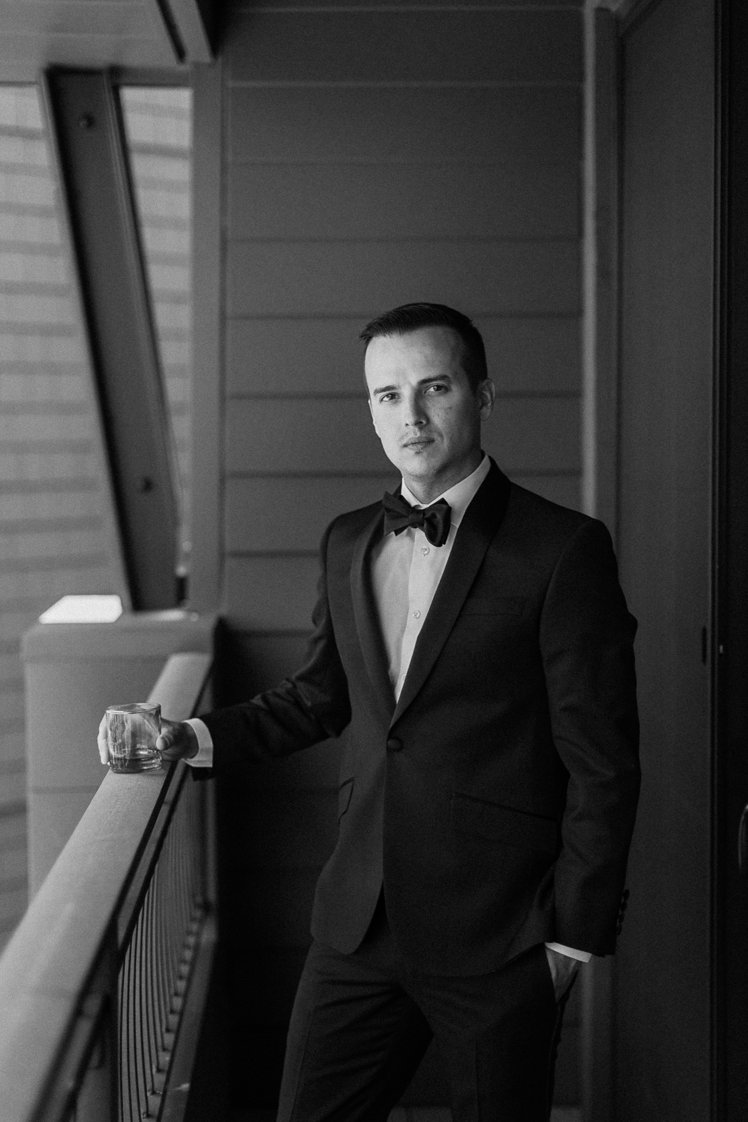 A groom in a tuxedo stands on a balcony over Lake Tahoe, holding a glass and looking seriously at the camera. Black-and-white photo.