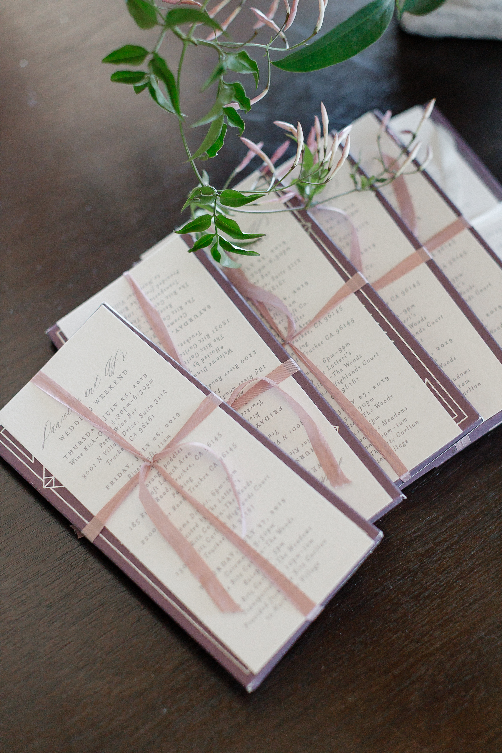 A stack of wedding invitations tied with pink ribbon and green sprigs sits on a dark wooden table, ready for a Lake Tahoe event.