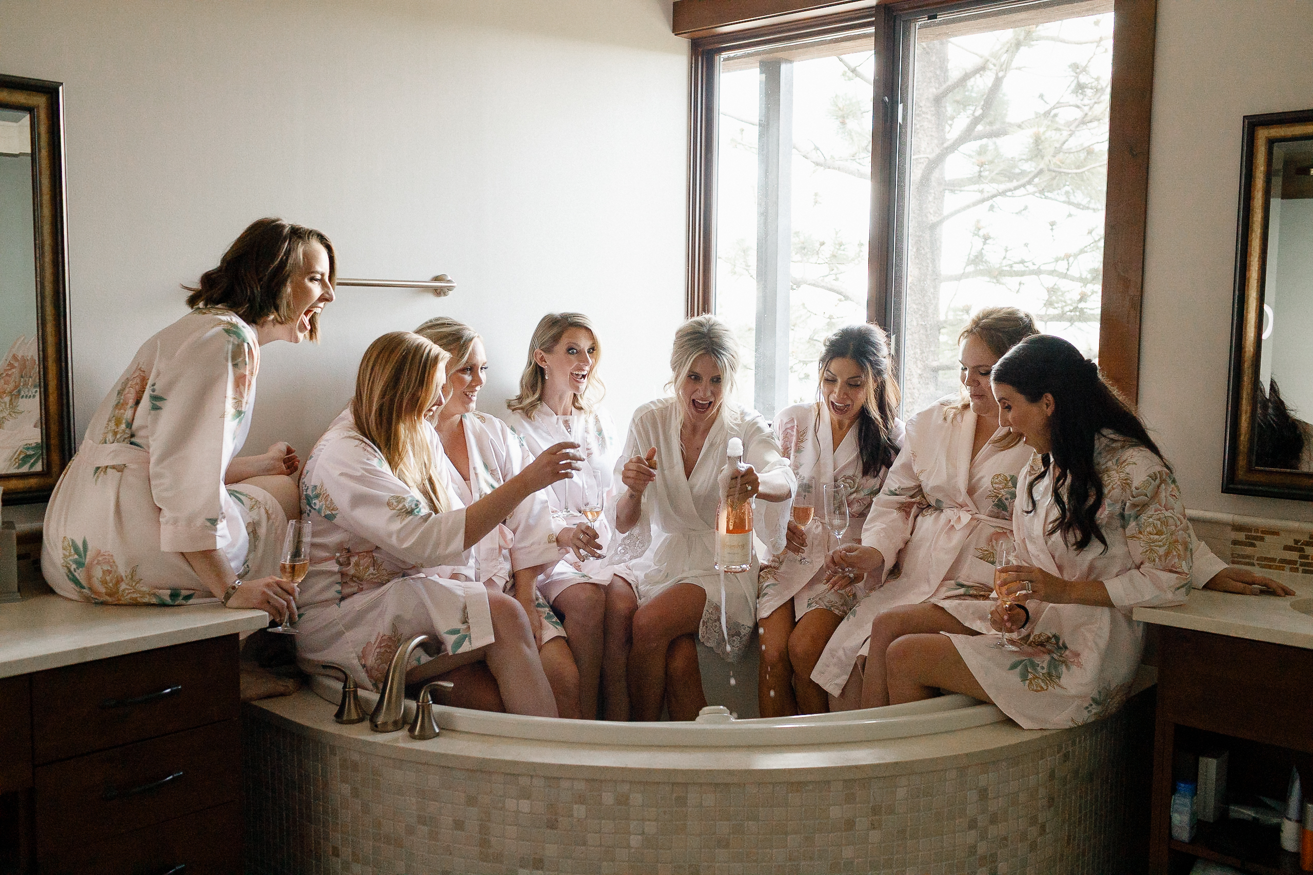 Seven women in floral robes gather by a bathtub at the Ritz-Carlton Lake Tahoe, laughing as one opens sparkling wine before a wedding.