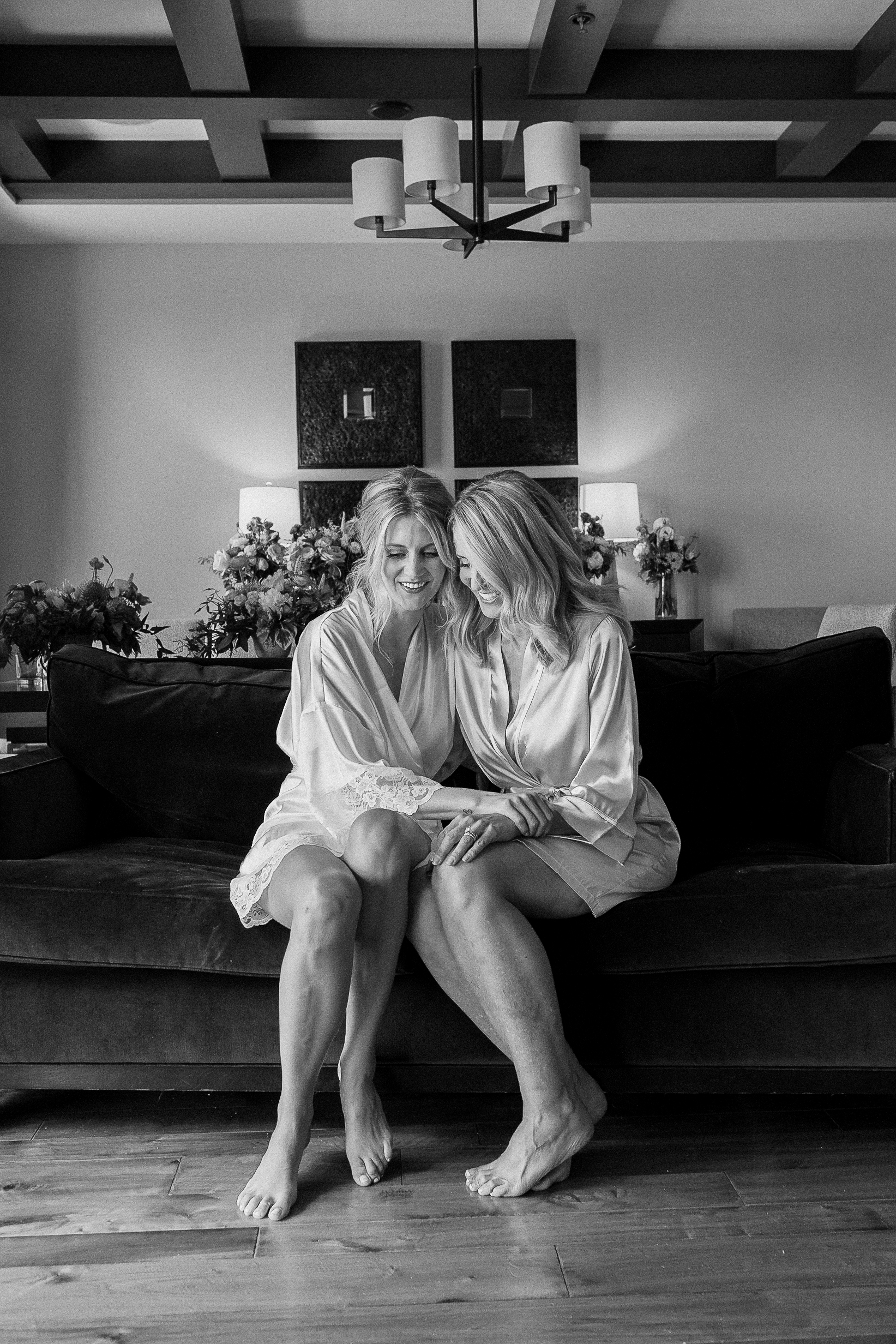 Two women in matching robes sit together on a couch, smiling and holding hands in a warm, flower-filled room before a Tahoe wedding.