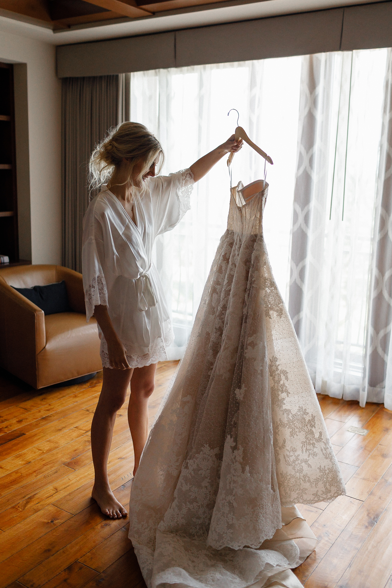 A woman in a robe stands by a window holding a wedding gown with sunlight streaming in on a quiet Lake Tahoe morning.