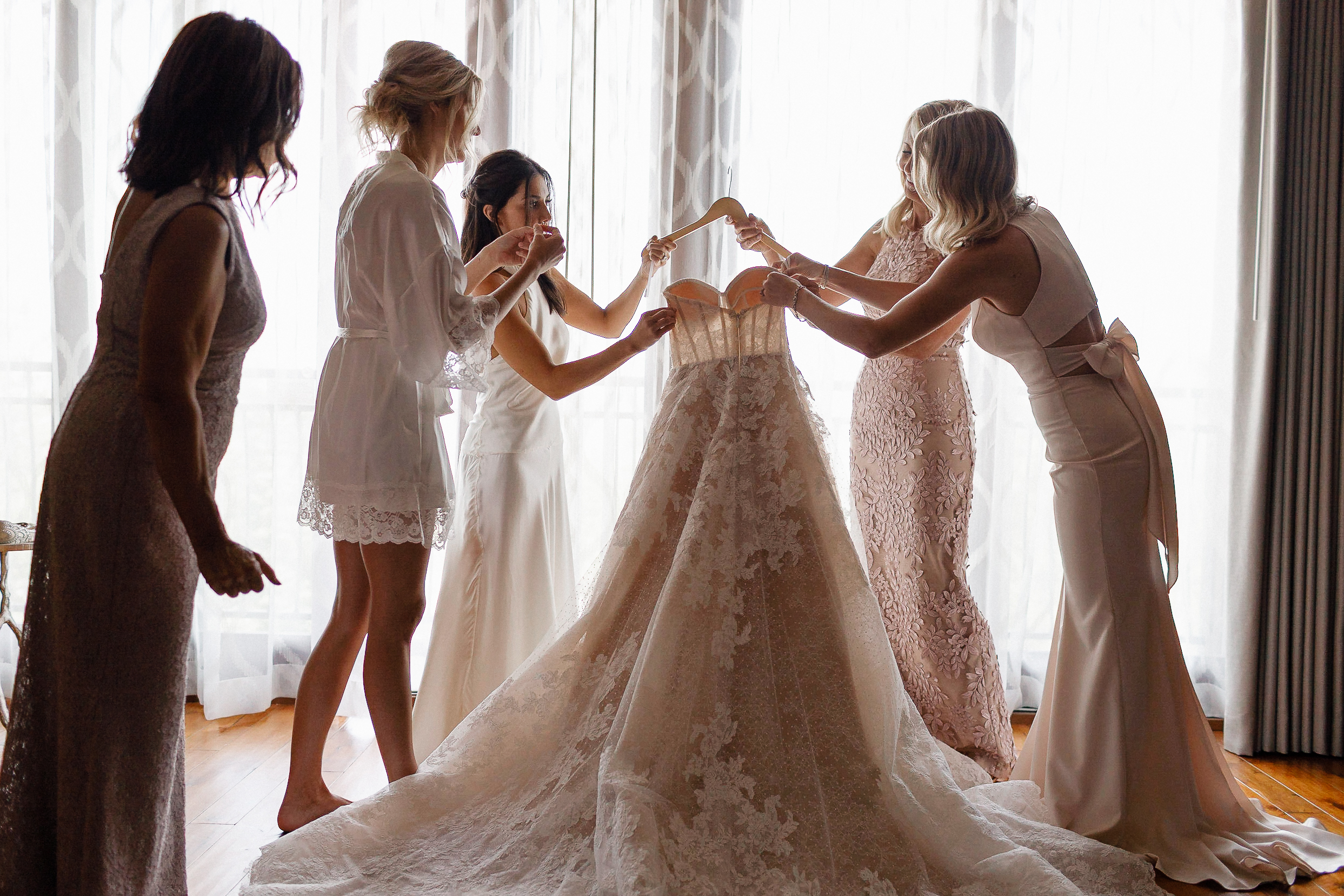 Four women help a bride hold up an ornate lace wedding dress on a hanger in a softly lit room at the Ritz-Carlton, Lake Tahoe.