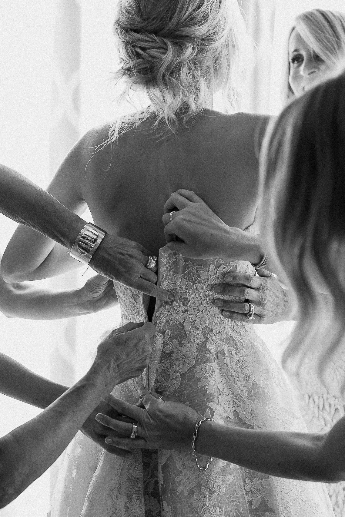 Several women help zip up a bride’s lace dress at the Ritz-Carlton; focus is on hands and the bride’s back, shot in black and white.