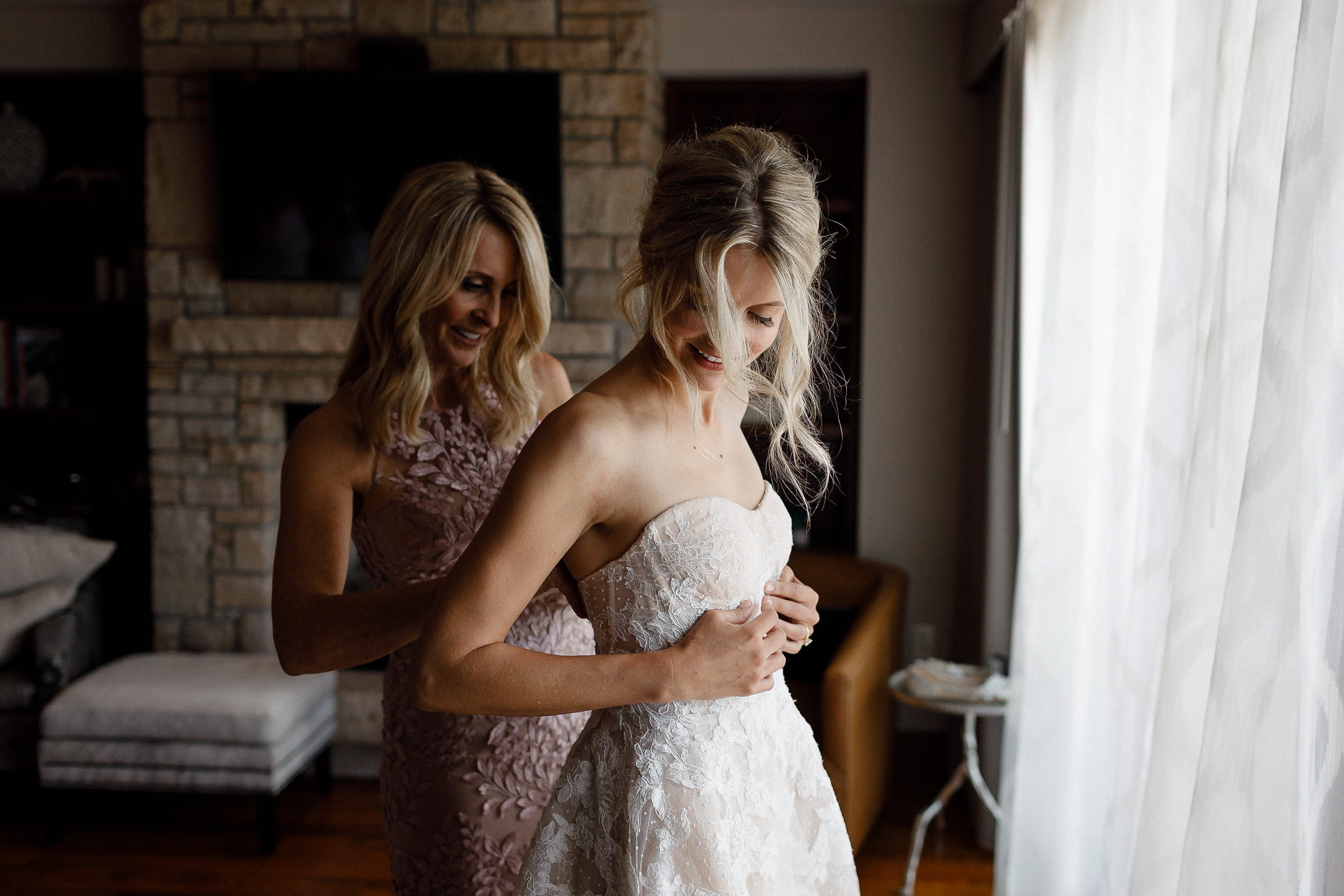 A bride in a strapless white gown smiles as a woman in a blush lace dress helps her by a window in a cozy Lake Tahoe room.