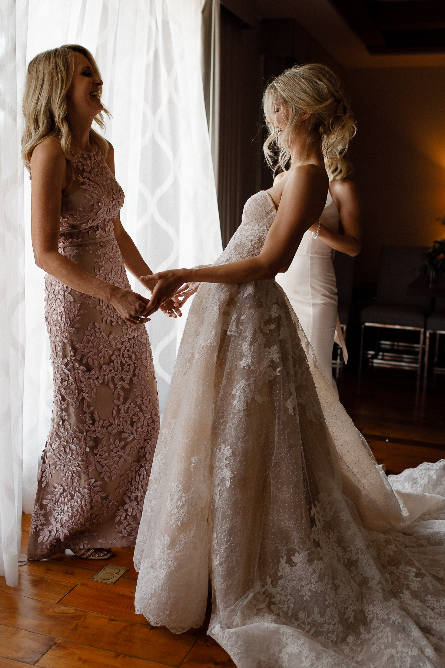 Two women hold hands and smile by a window at the Ritz-Carlton, Lake Tahoe, dressed in lace wedding and blush pink dresses.