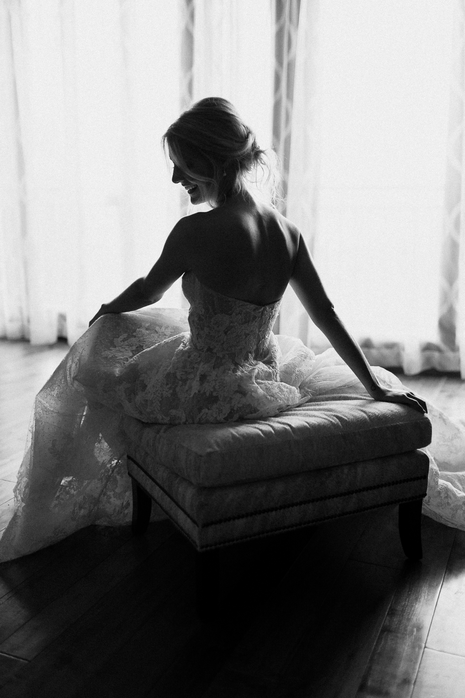 Woman in a strapless lace wedding dress sits sideways on a bench at the Ritz-Carlton Lake Tahoe, outlined by soft window light.