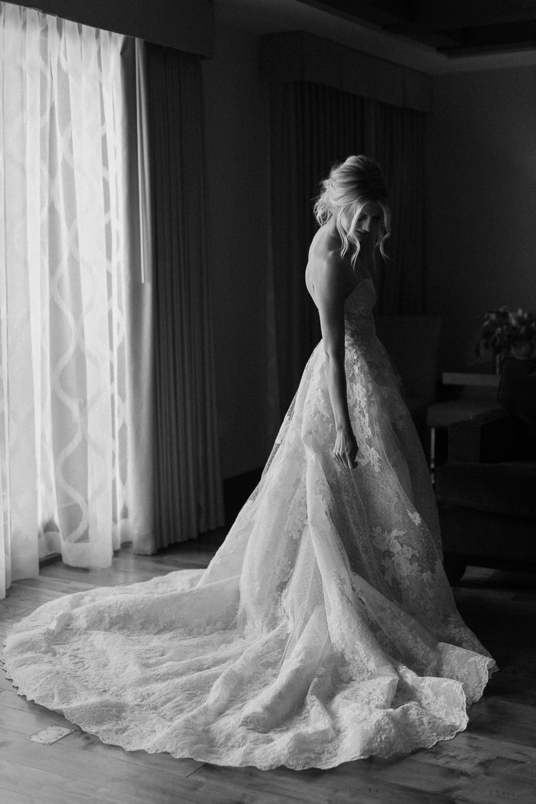 Bride in strapless lace Watters dress with long train stands by large window at Ritz-Carlton, lit by soft natural light.