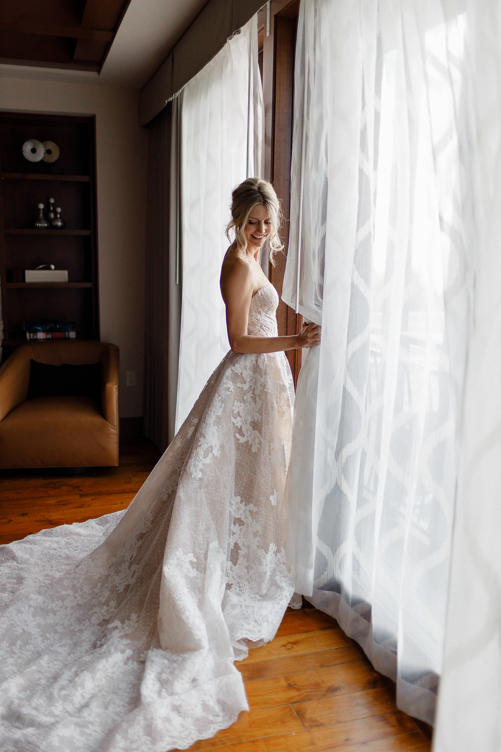 Bride in lace wedding dress stands by window with sheer curtains at Ritz-Carlton Lake Tahoe. Sunlight highlights her long train.