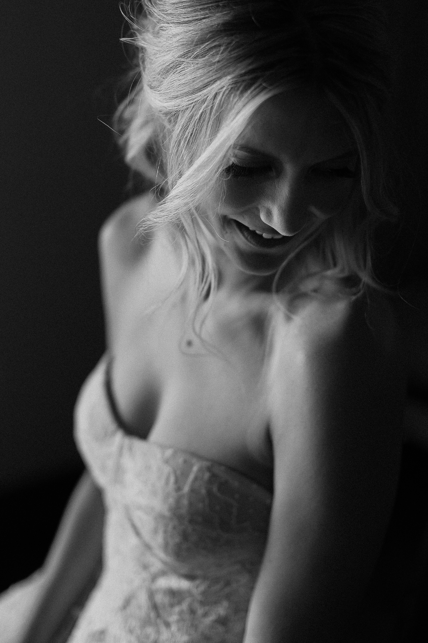 Black and white photo of a woman with wavy blonde updo, smiling down in a strapless lace wedding dress. Light highlights her face.