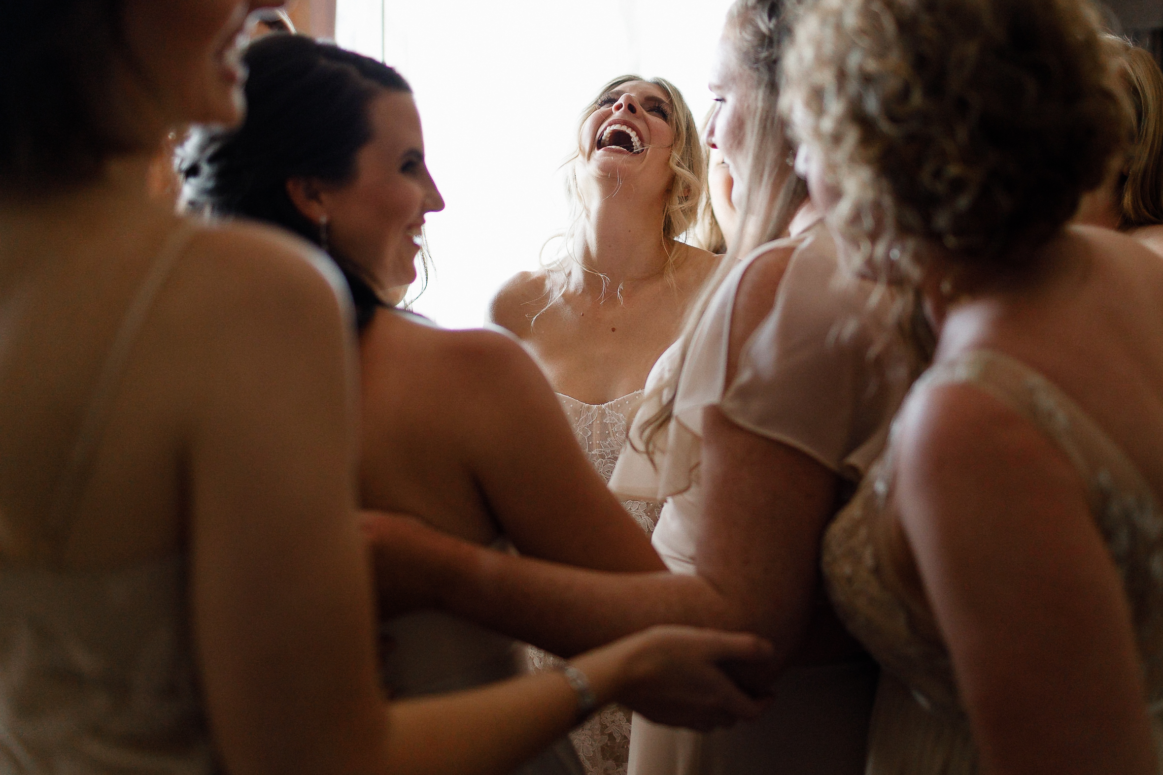 A bride in a white dress laughs with bridesmaids in light dresses, all enjoying a joyful, close moment together indoors.