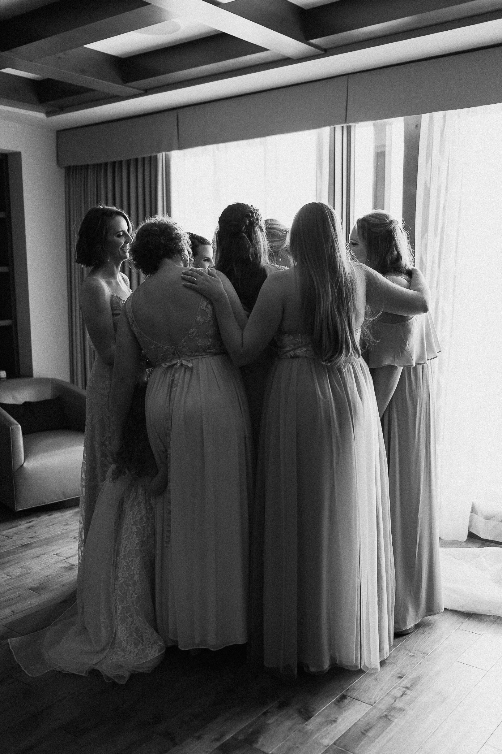 Women in dresses huddle and share a joyful moment by a window at the Ritz-Carlton before a Lake Tahoe wedding; black and white photo.