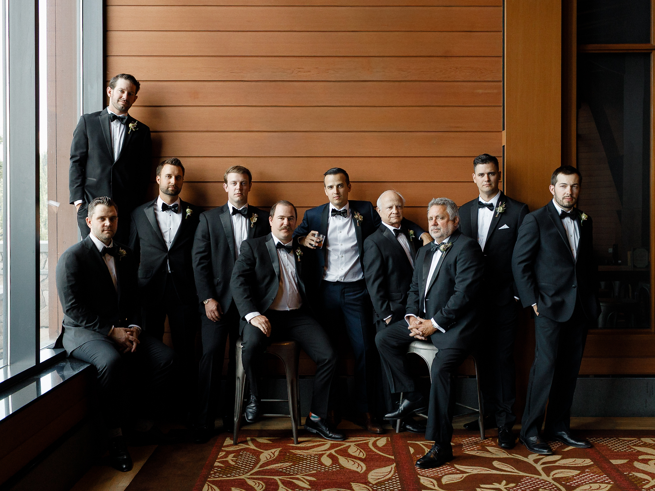 Eleven men in black tuxedos pose indoors by a window and wood wall at the Ritz-Carlton Lake Tahoe, standing and seated.
