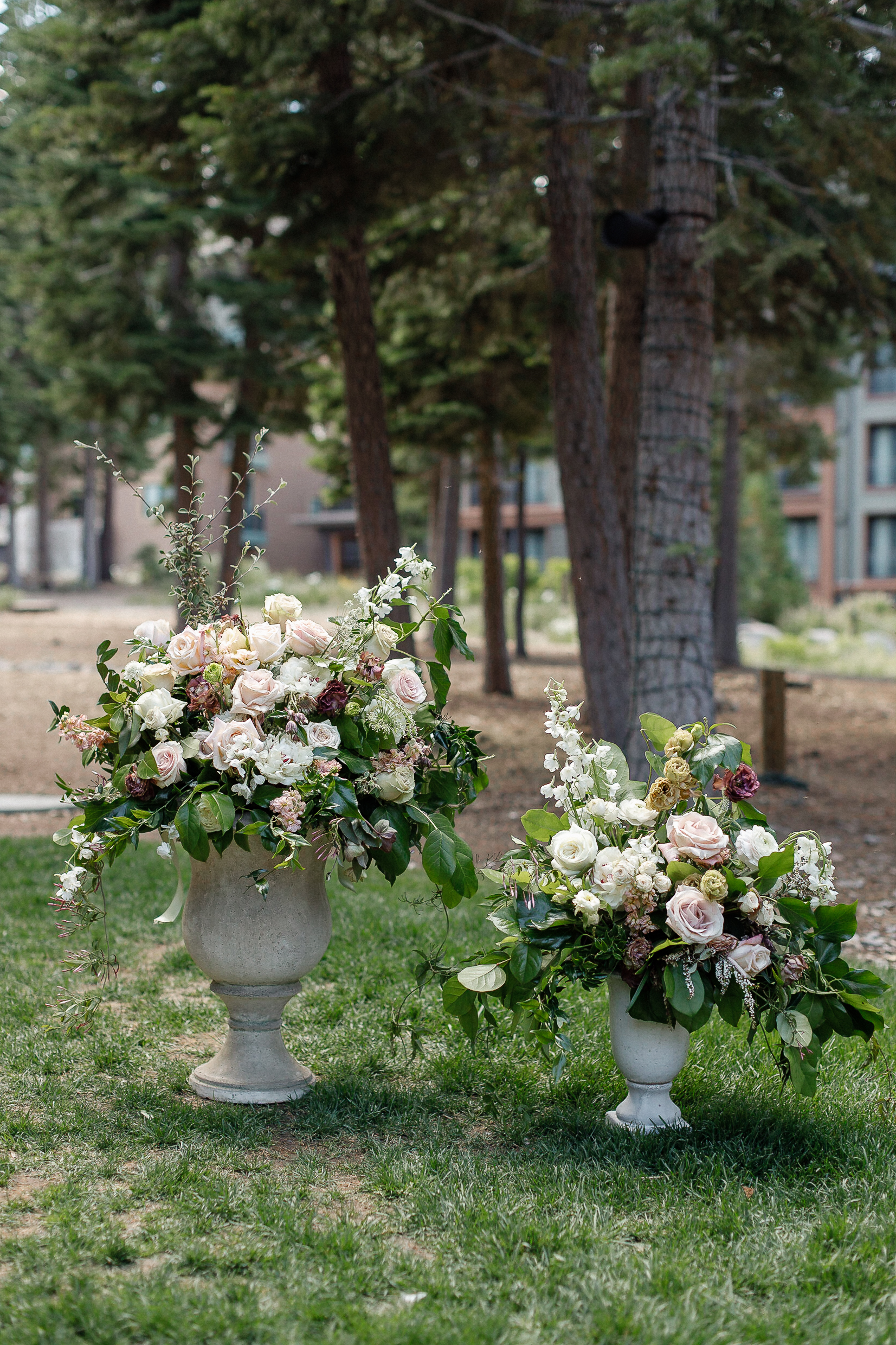 Two large stone urns with white, blush, and pink roses and greenery sit on grass, with trees and Ritz-Carlton buildings behind.