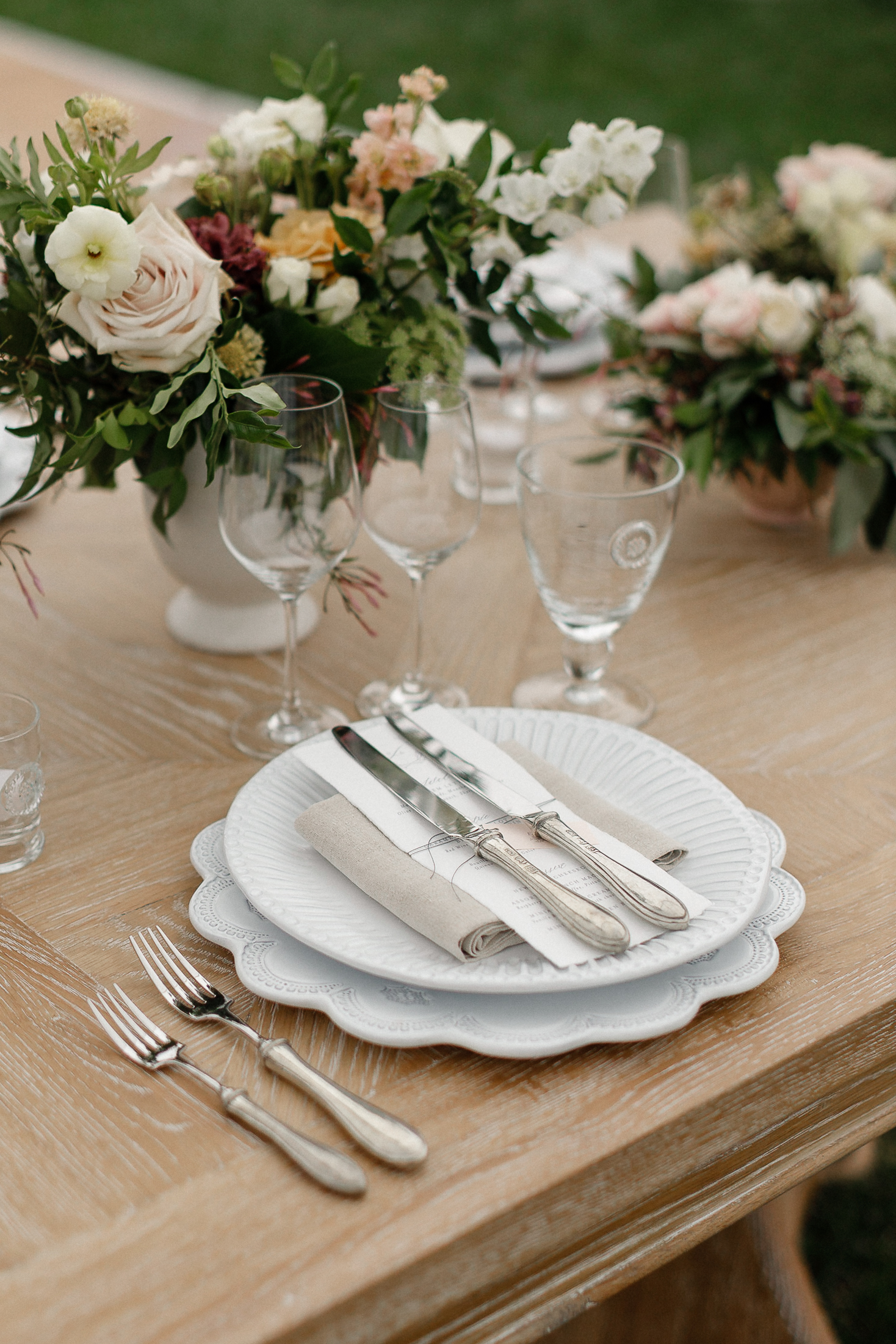 Table set with layered white plates, rolled napkin and cutlery, wine glasses, and pastel floral centerpieces on wooden table.