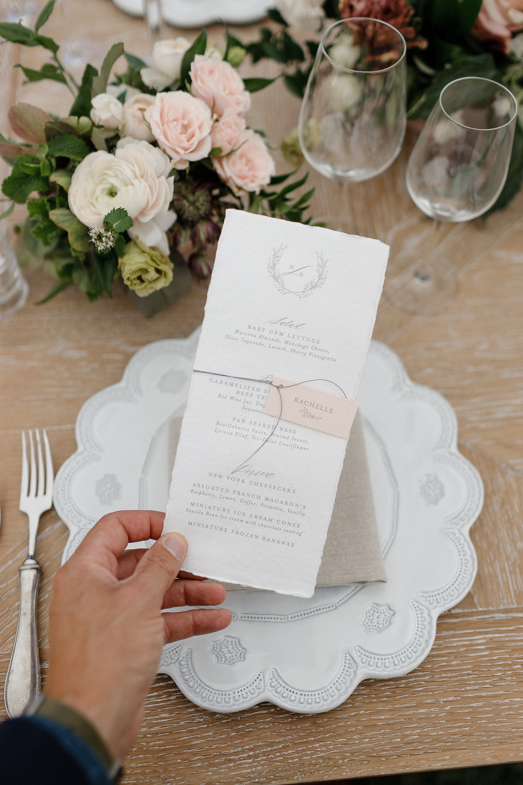 Hand holding a Ritz-Carlton wedding menu with floral accents on a plate, with wine glasses, silverware, and pink-white flowers.