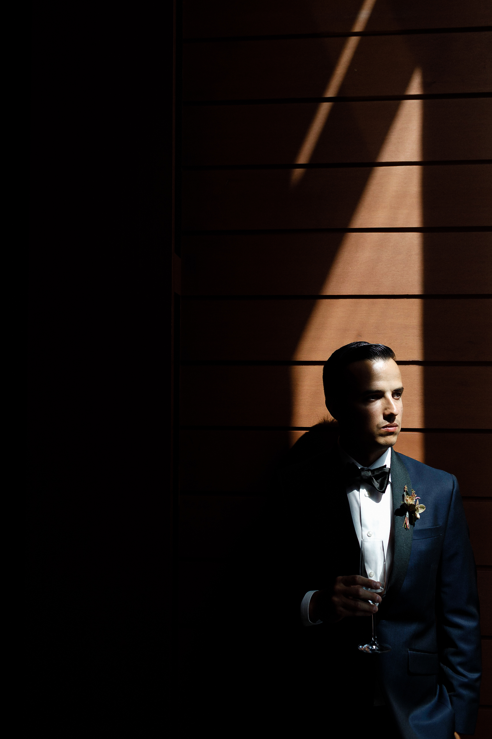 A man in a tuxedo stands by a wooden wall at the Ritz-Carlton, holding a glass, partly lit by sunlight. The rest is in shadow.