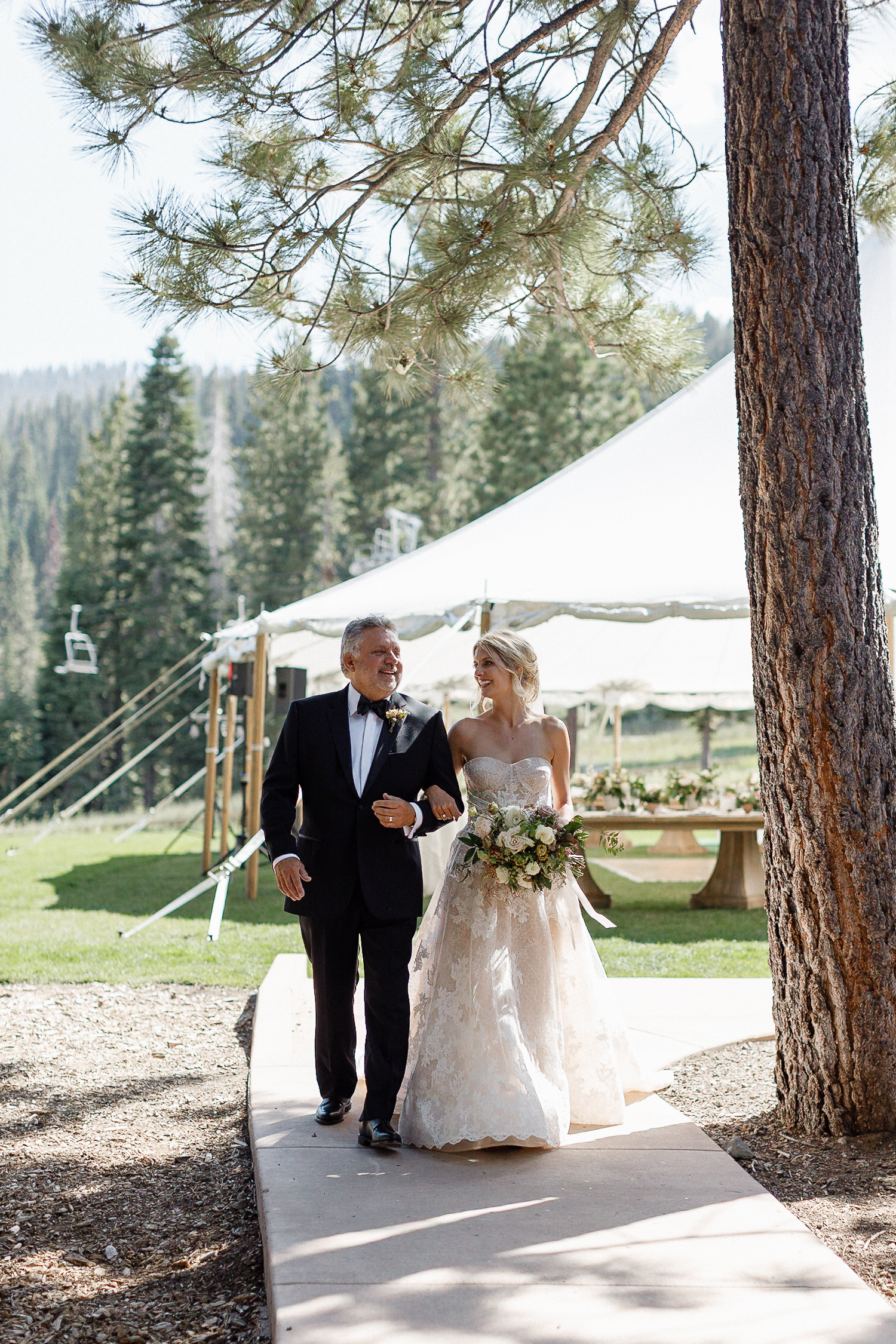 Bride in strapless gown holding bouquet walks with older man in tuxedo at Ritz-Carlton wedding; tent, tables, pines behind.