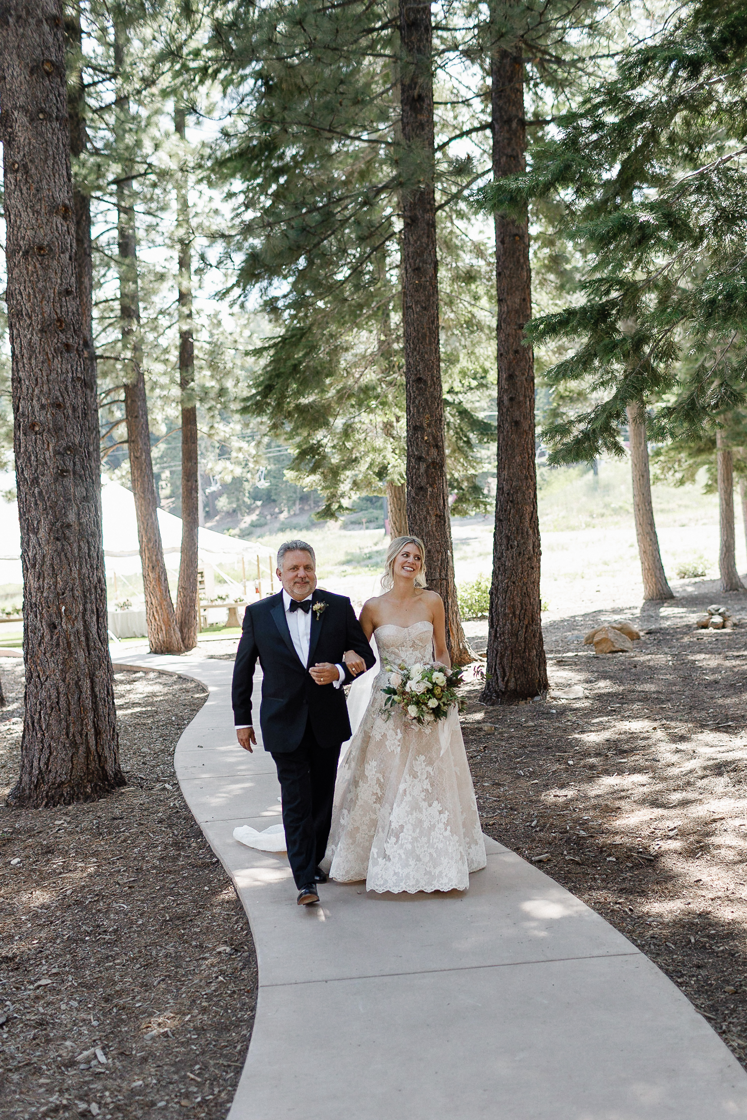 Bride in strapless gown walks arm-in-arm with her father along a sunlit, forested path near Lake Tahoe, holding a bouquet.