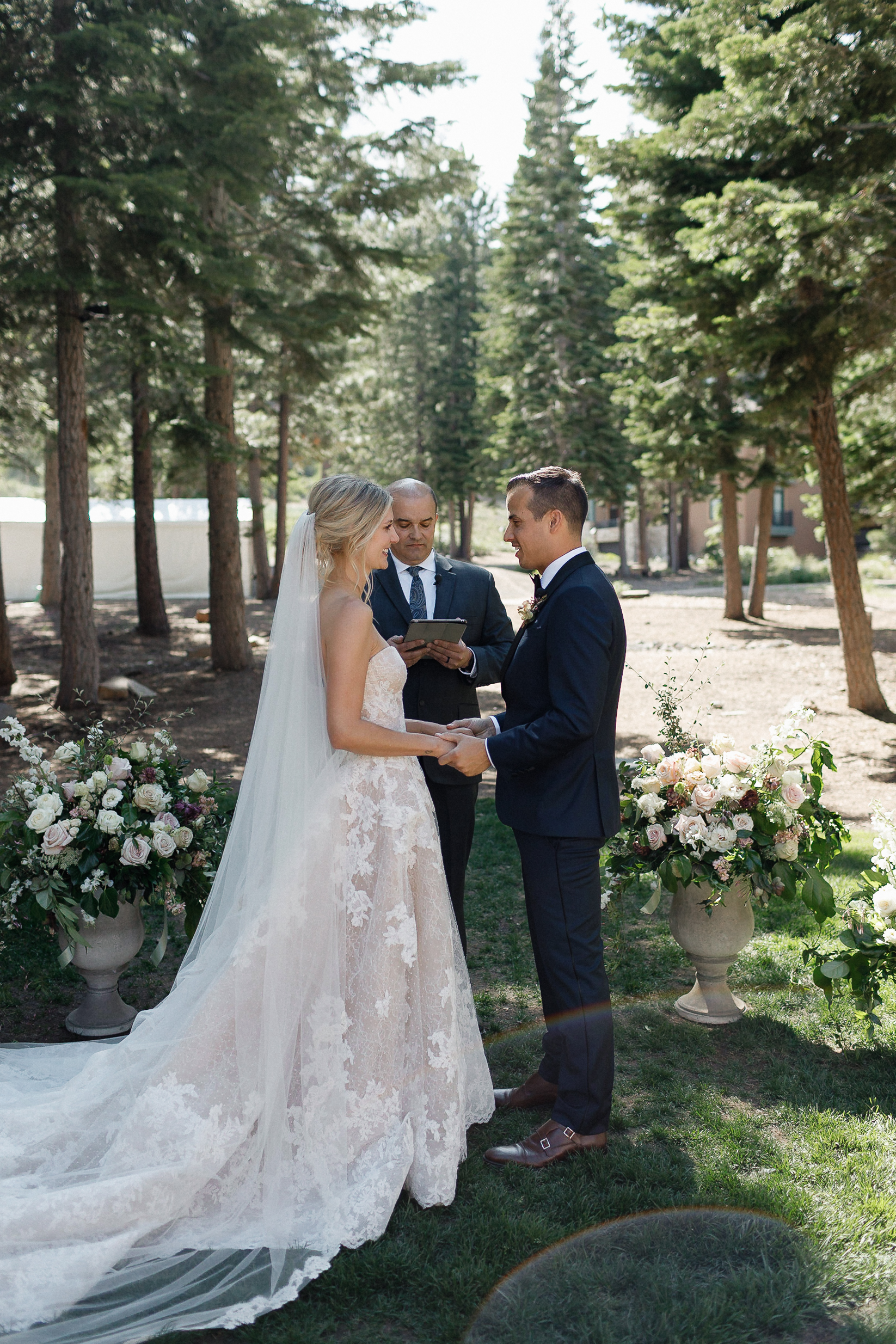 Bride and groom hold hands and exchange vows in a forest near Lake Tahoe, with an officiant and floral arrangements beside them.
