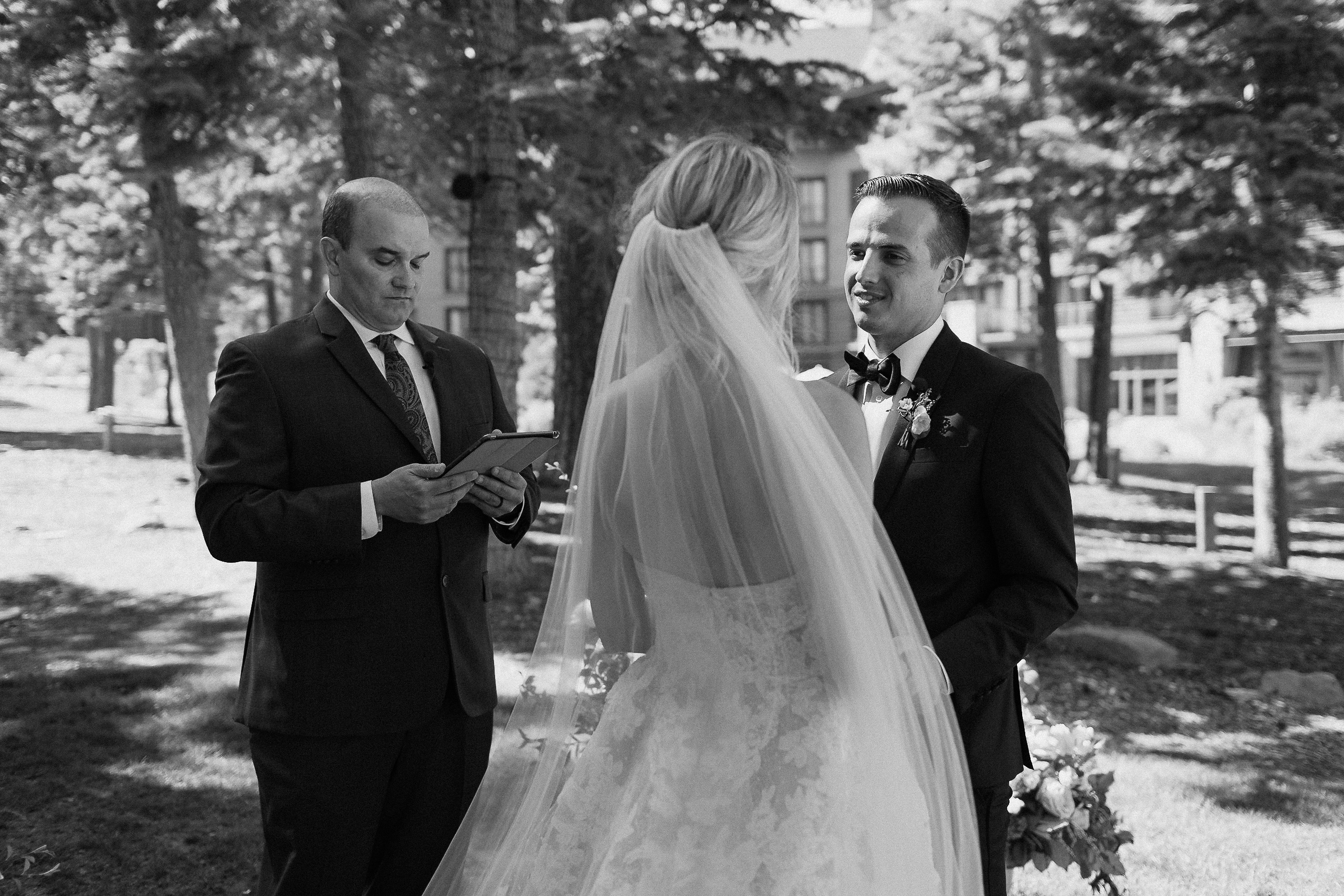 Bride and groom smile at each other outdoors as an officiant reads from a tablet; tall trees and sunlight fill the Ritz-Carlton background.