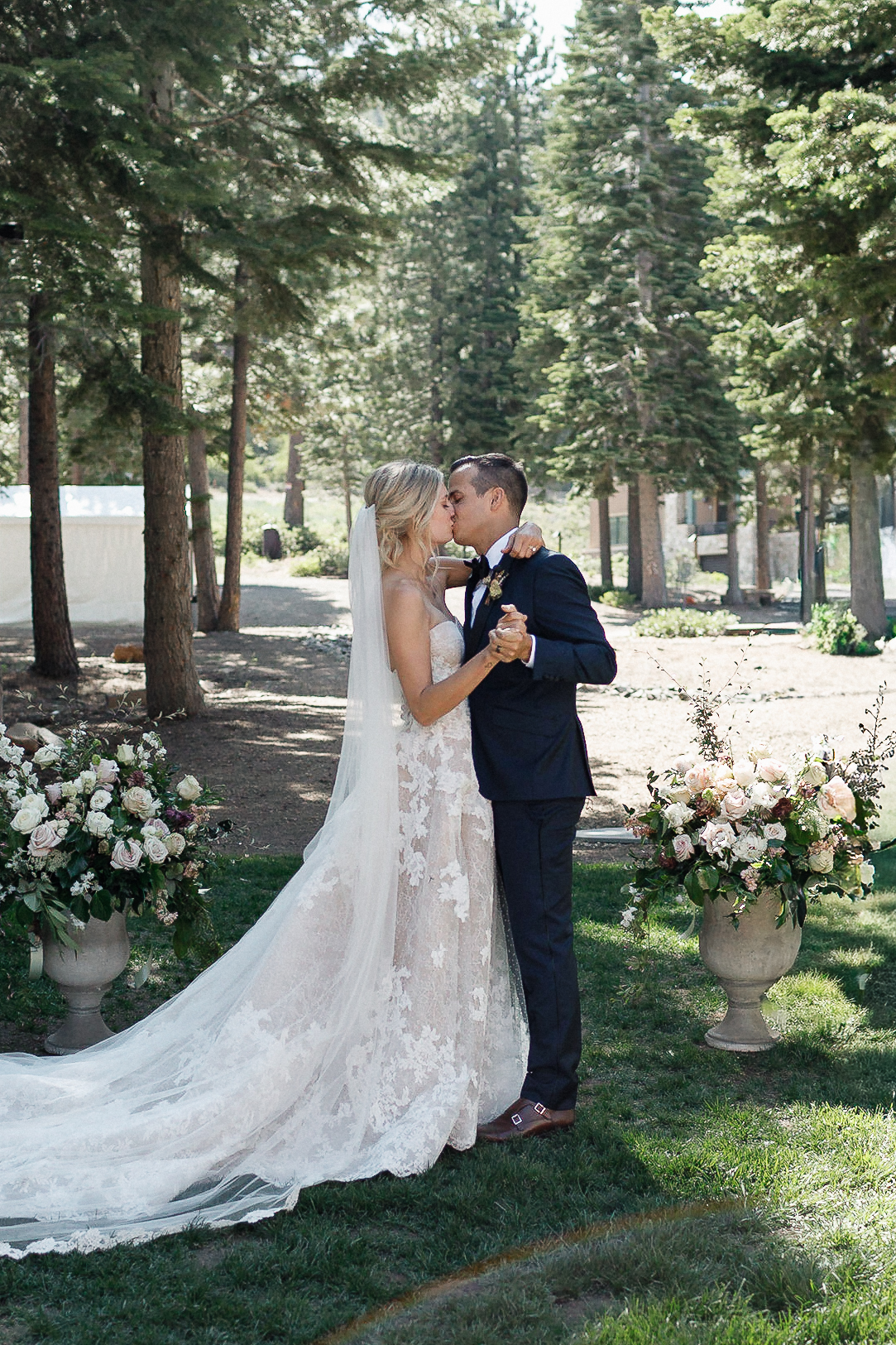 Bride and groom share first kiss outdoors at Lake Tahoe, between two floral arrangements. Bride in lace dress; groom in dark suit.