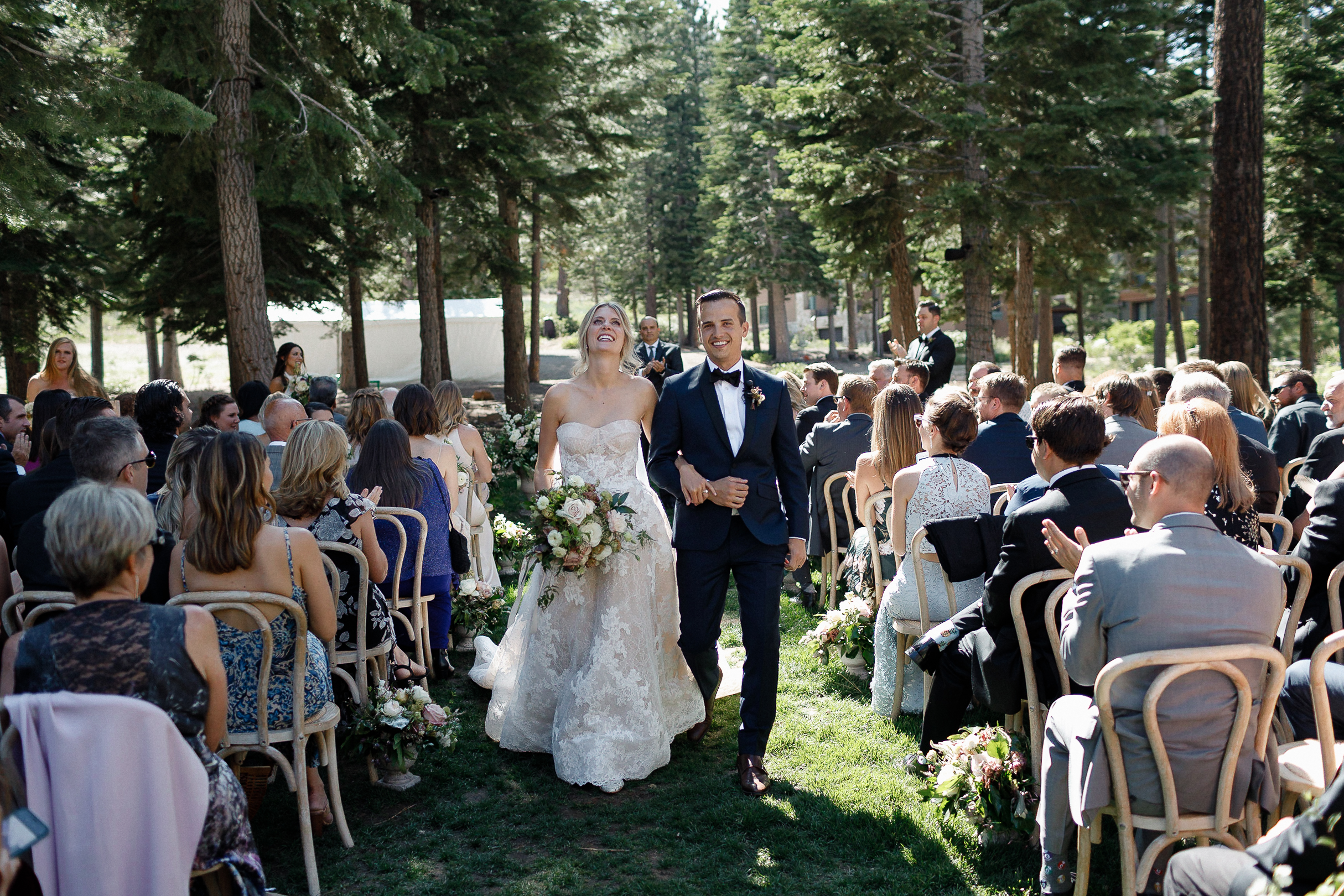 A smiling bride and groom hold hands walking down an outdoor aisle at a sunny forest wedding, guests seated among tall trees.