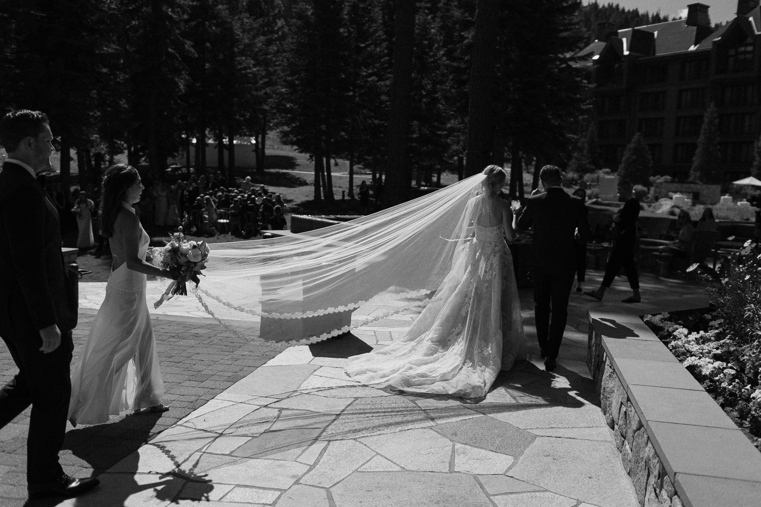 Bride and groom walk outdoors at Ritz-Carlton Lake Tahoe, with bridal party behind; guests stand near tall trees and buildings.