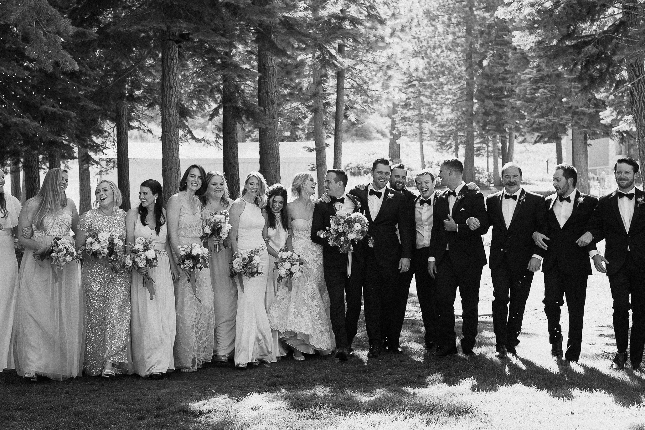 Large wedding party walks together outdoors by Lake Tahoe, bridesmaids in light dresses and groomsmen in tuxedos, all smiling.