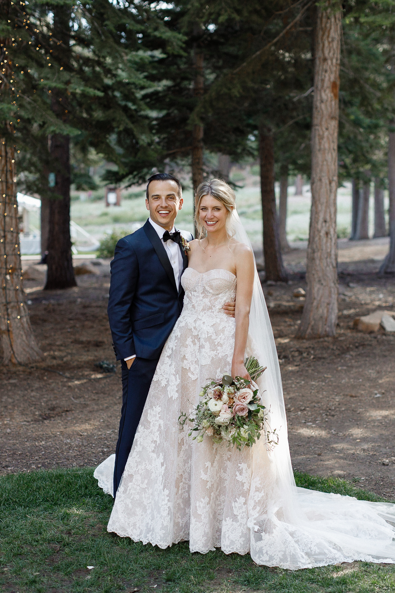 Bride in white lace gown and veil stands with groom in navy suit, both smiling outdoors by tall trees at Ritz-Carlton. She holds pale bouquet.