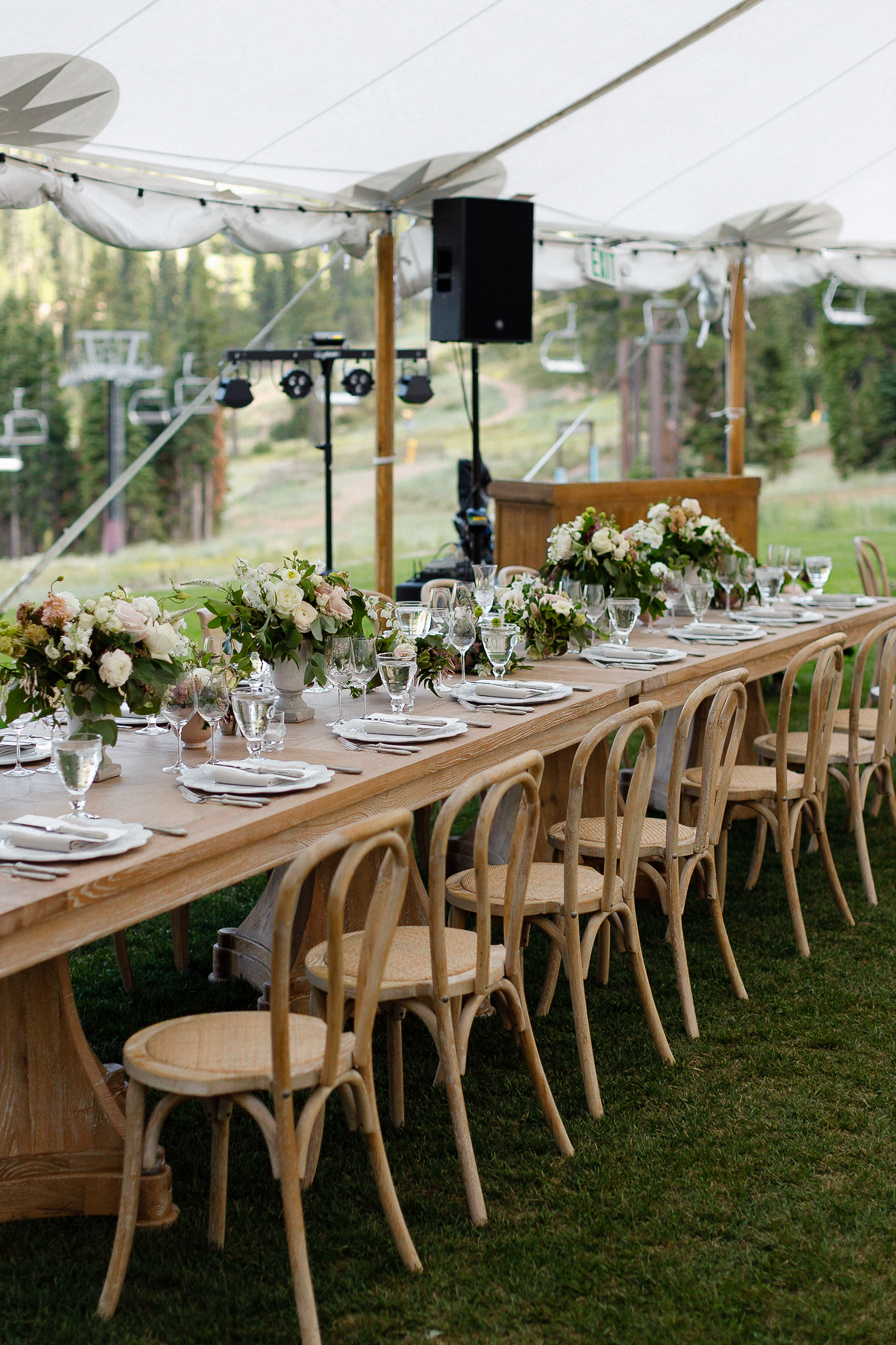Long wooden table set for a Lake Tahoe wedding under a white tent with floral centerpieces, glassware, chairs, and greenery.