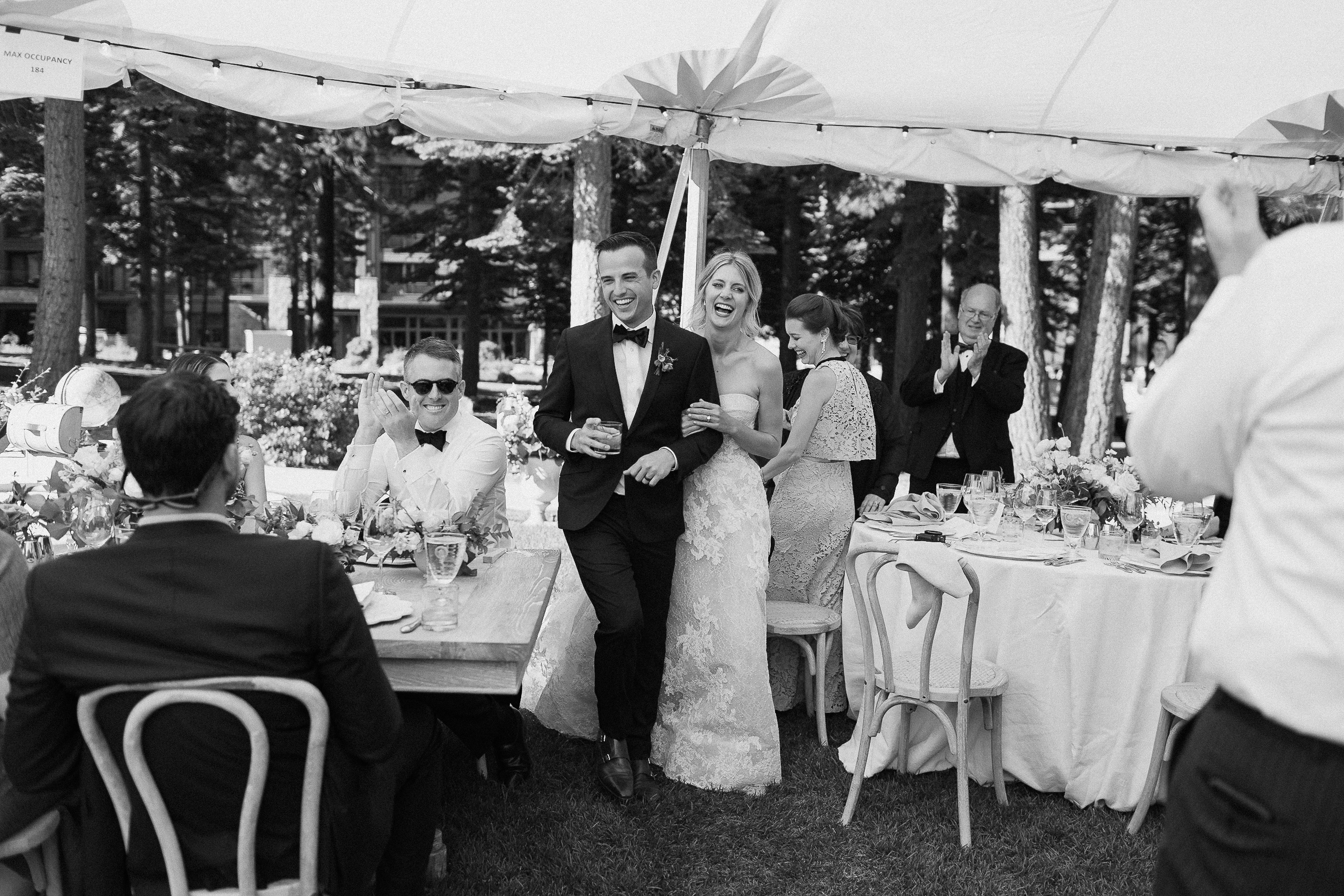 Bride and groom, smiling, enter a lively outdoor wedding under a tent at Ritz-Carlton Lake Tahoe as guests applaud and celebrate.
