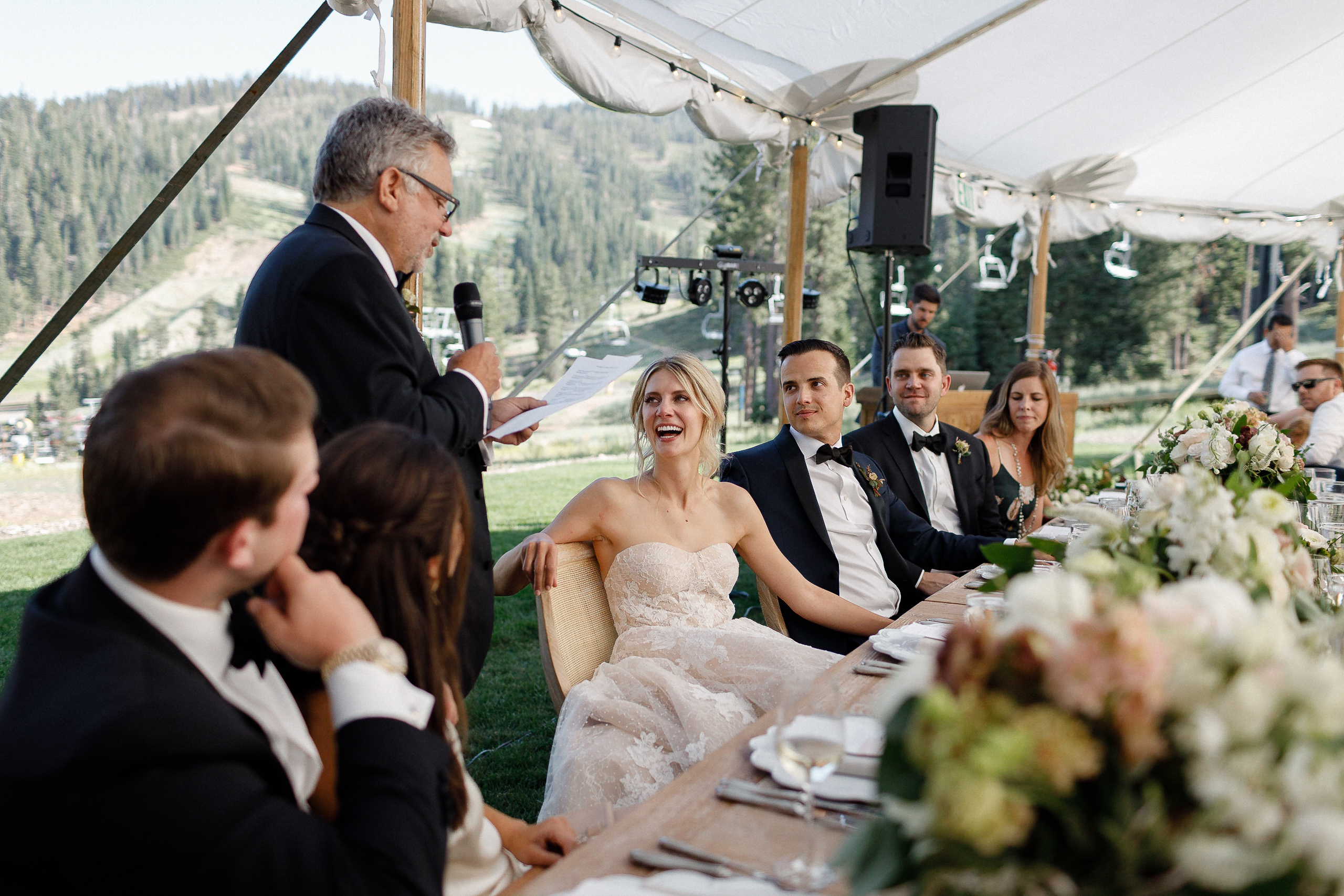 Bride in strapless gown smiles with groom and wedding party under tent as father gives toast; green mountains in background.
