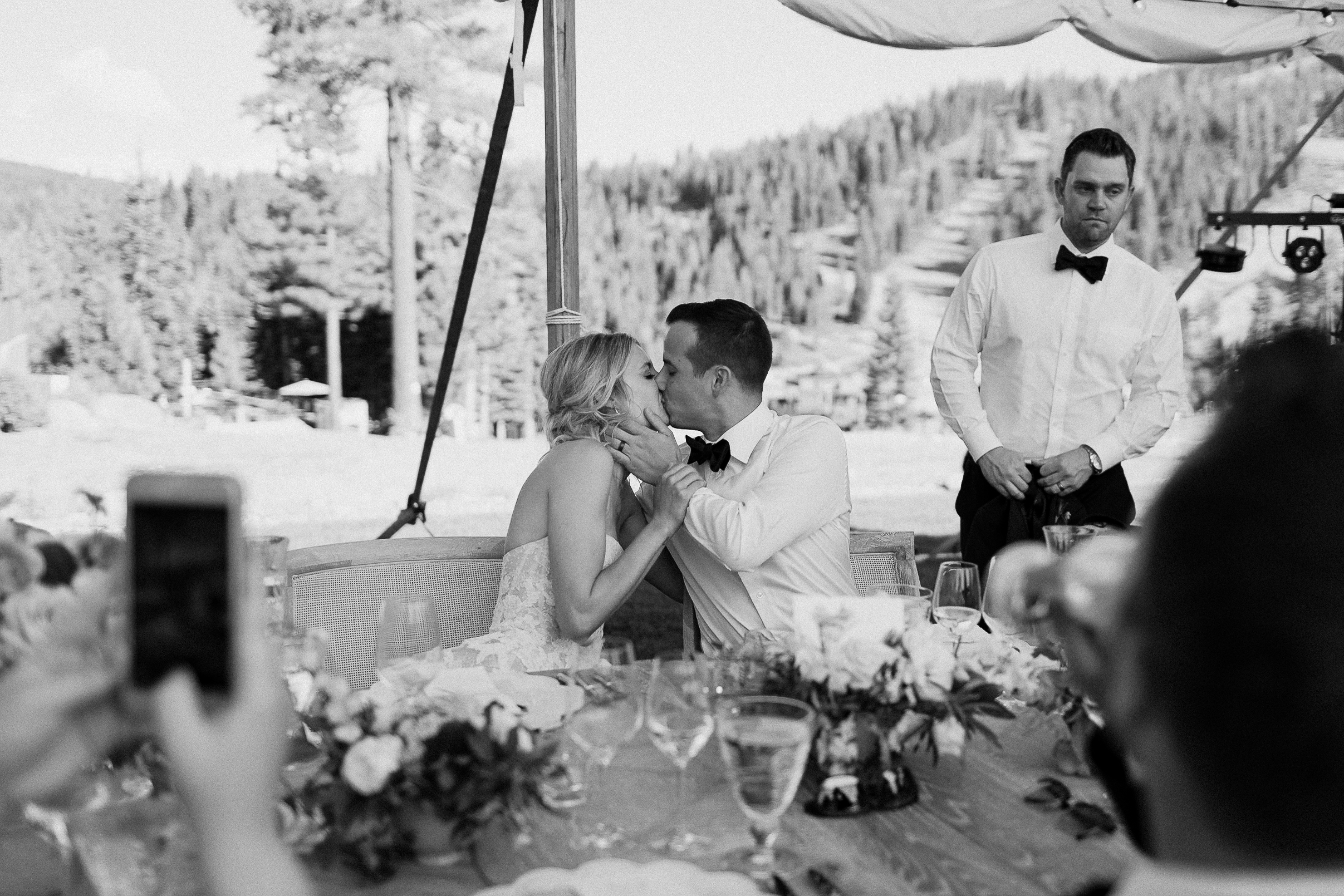 Newlyweds kiss at their Lake Tahoe reception table as guests watch and take photos, with Ritz-Carlton, trees, and mountains behind.