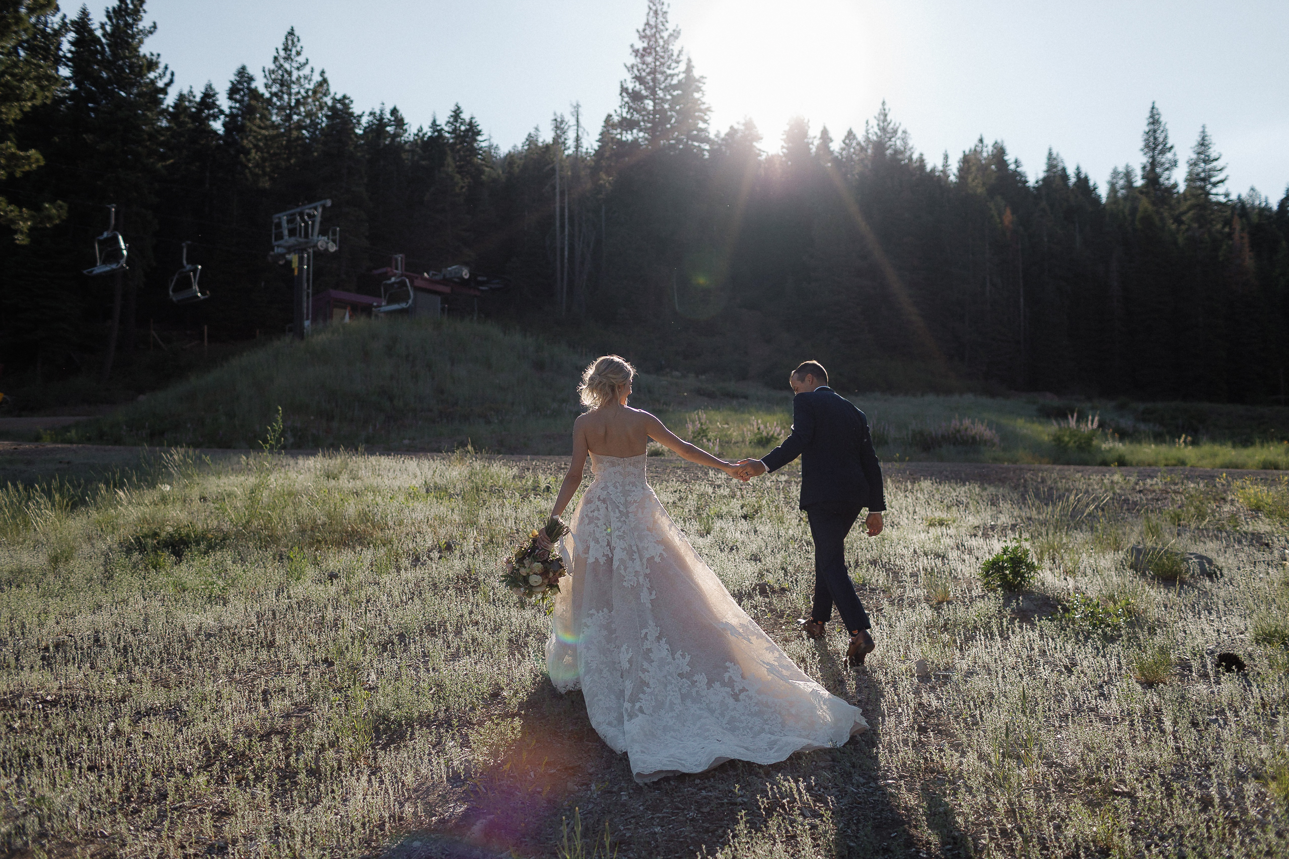 Bride and groom walk hand in hand through sunlit grass near Lake Tahoe, with trees, mountains, and ski lifts in the background.
