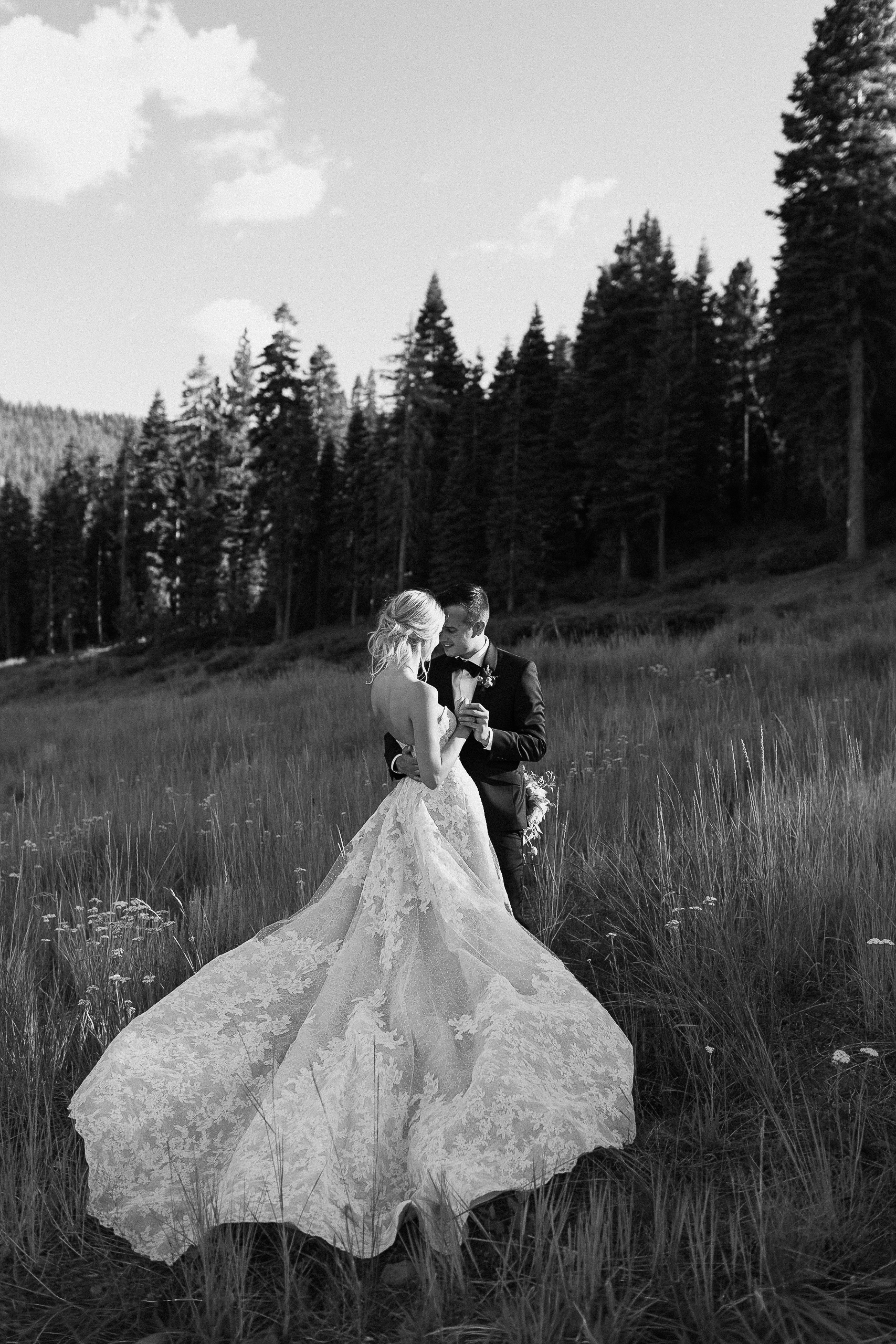 Bride in lace gown stands in tall grass with groom in suit, surrounded by pine trees and mountains—a serene wedding portrait.