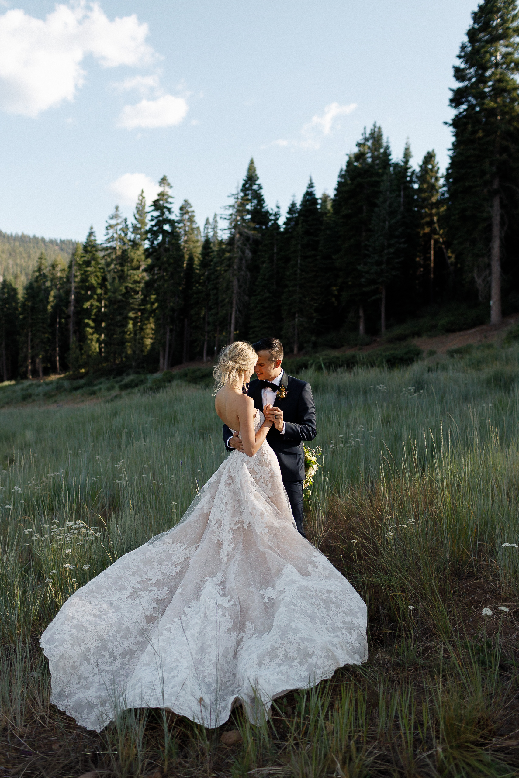 Bride in lace wedding gown and groom in dark suit gaze at each other in tall grass, with trees and mountains in the background.