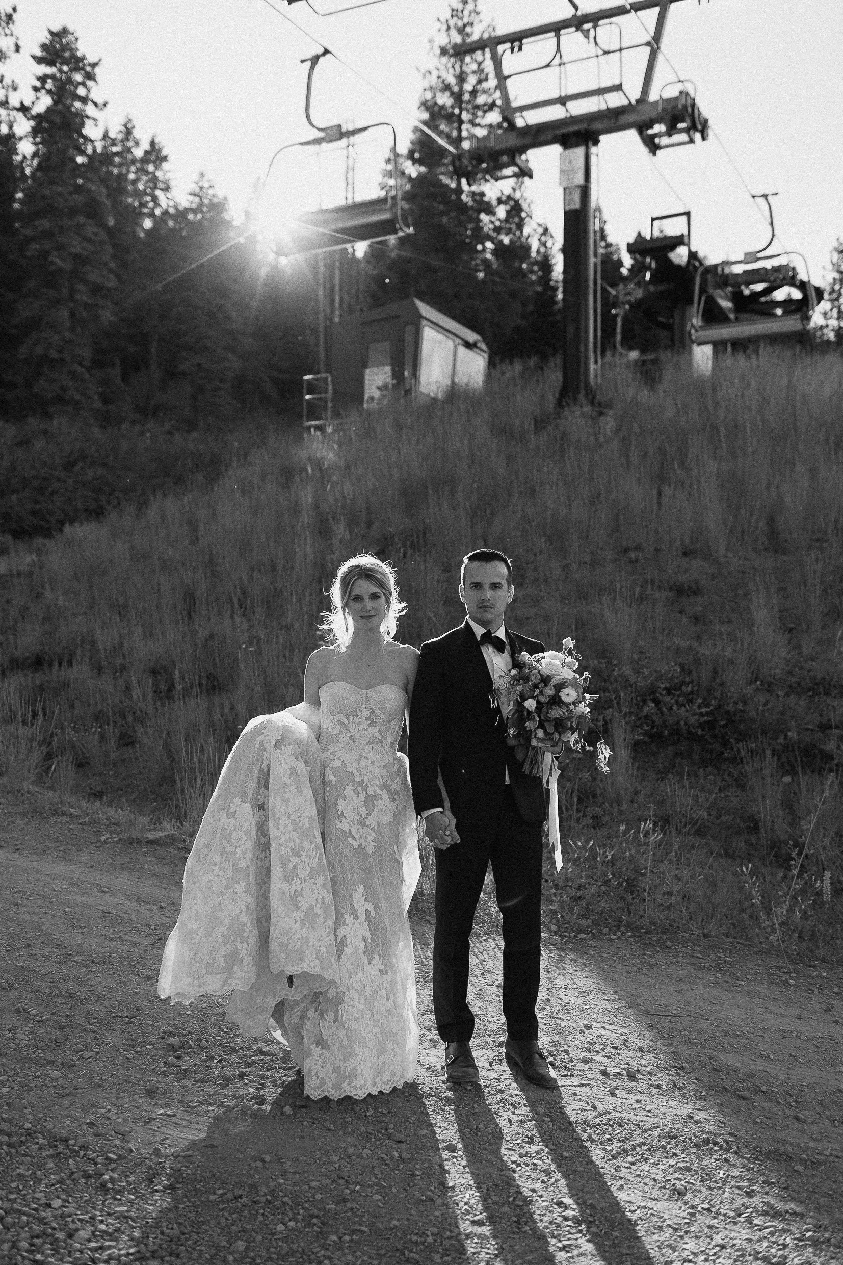 Bride in strapless lace gown and groom in tuxedo hold hands on a dirt path, with trees and sunlit ski lift in Lake Tahoe.