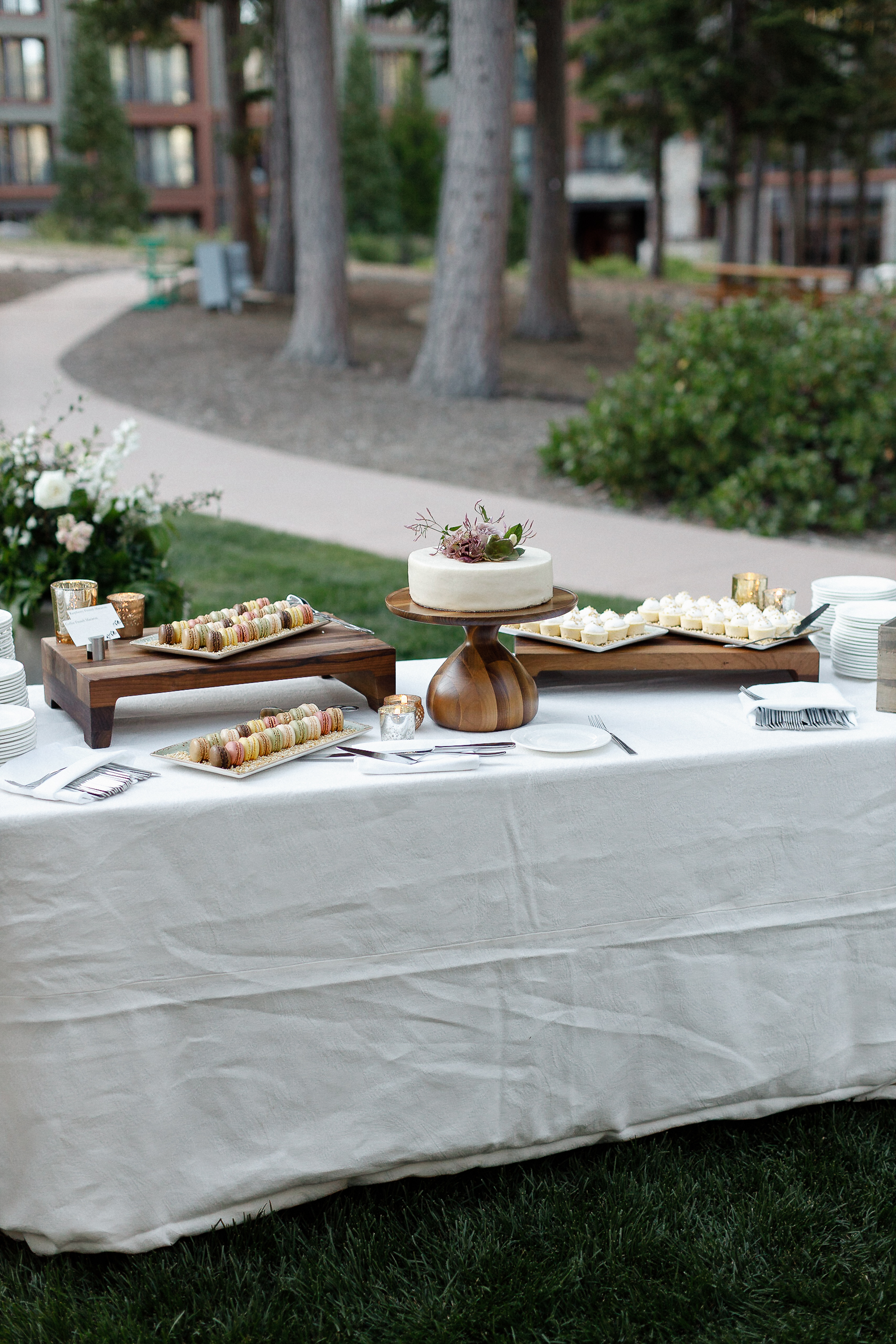Outdoor table with white tablecloth holds macarons, cookies, and a cake on a wooden stand; trees and buildings in background.