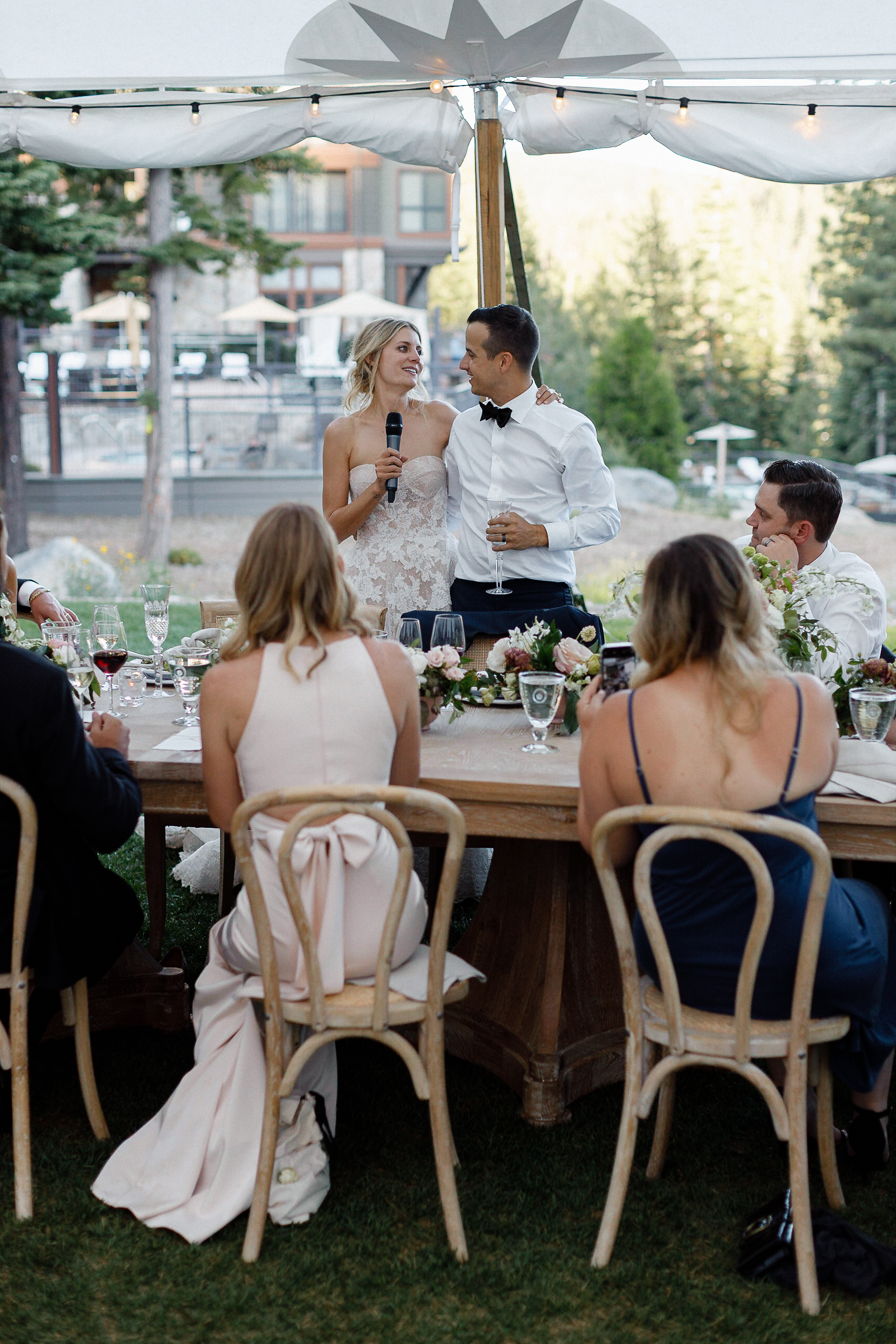 Bride with microphone and groom smile together at their Lake Tahoe wedding, guests seated at flower-decorated wooden tables.