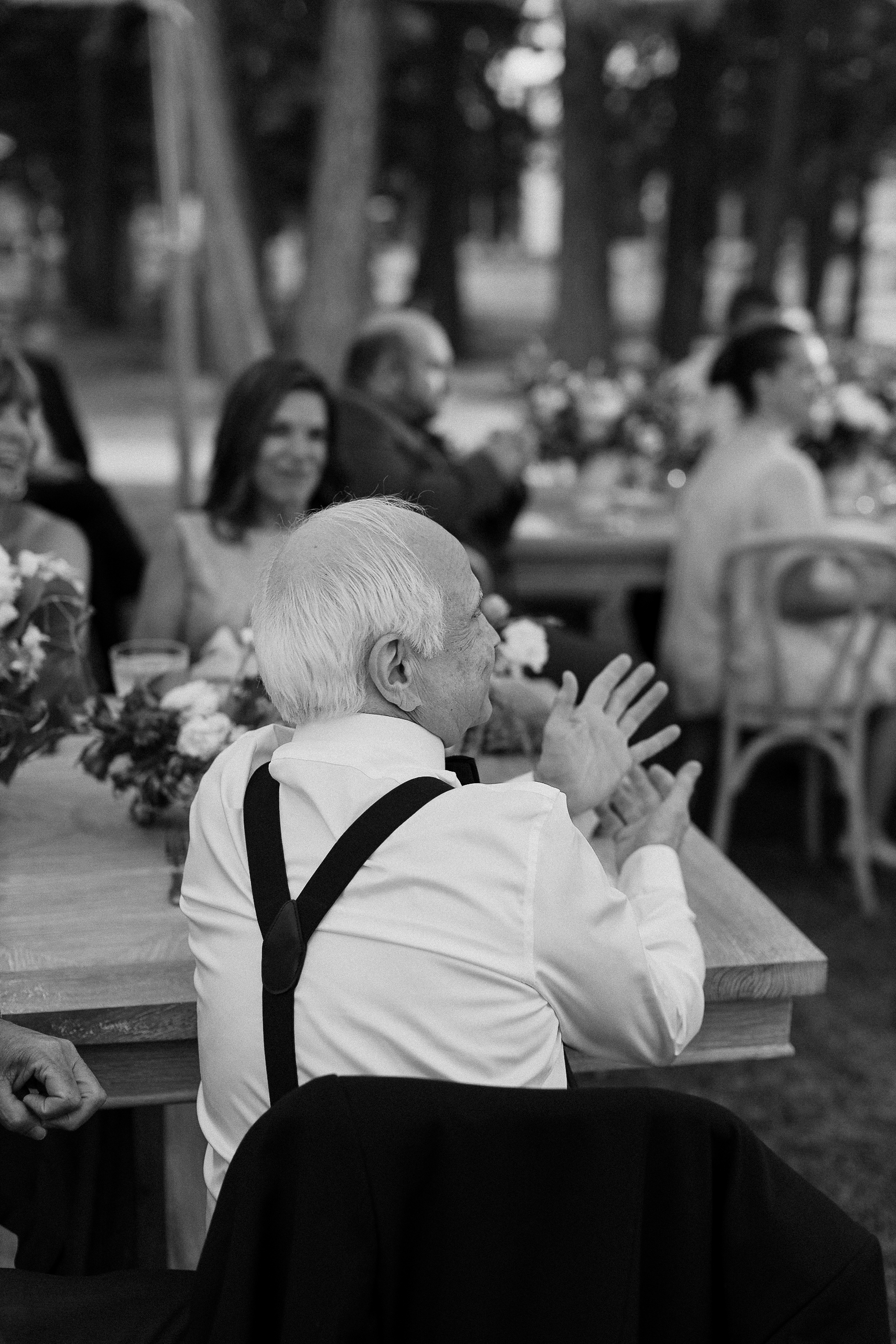 Elderly man with white hair, white shirt, and suspenders sits outside at a wooden table, smiling and clapping among happy guests.
