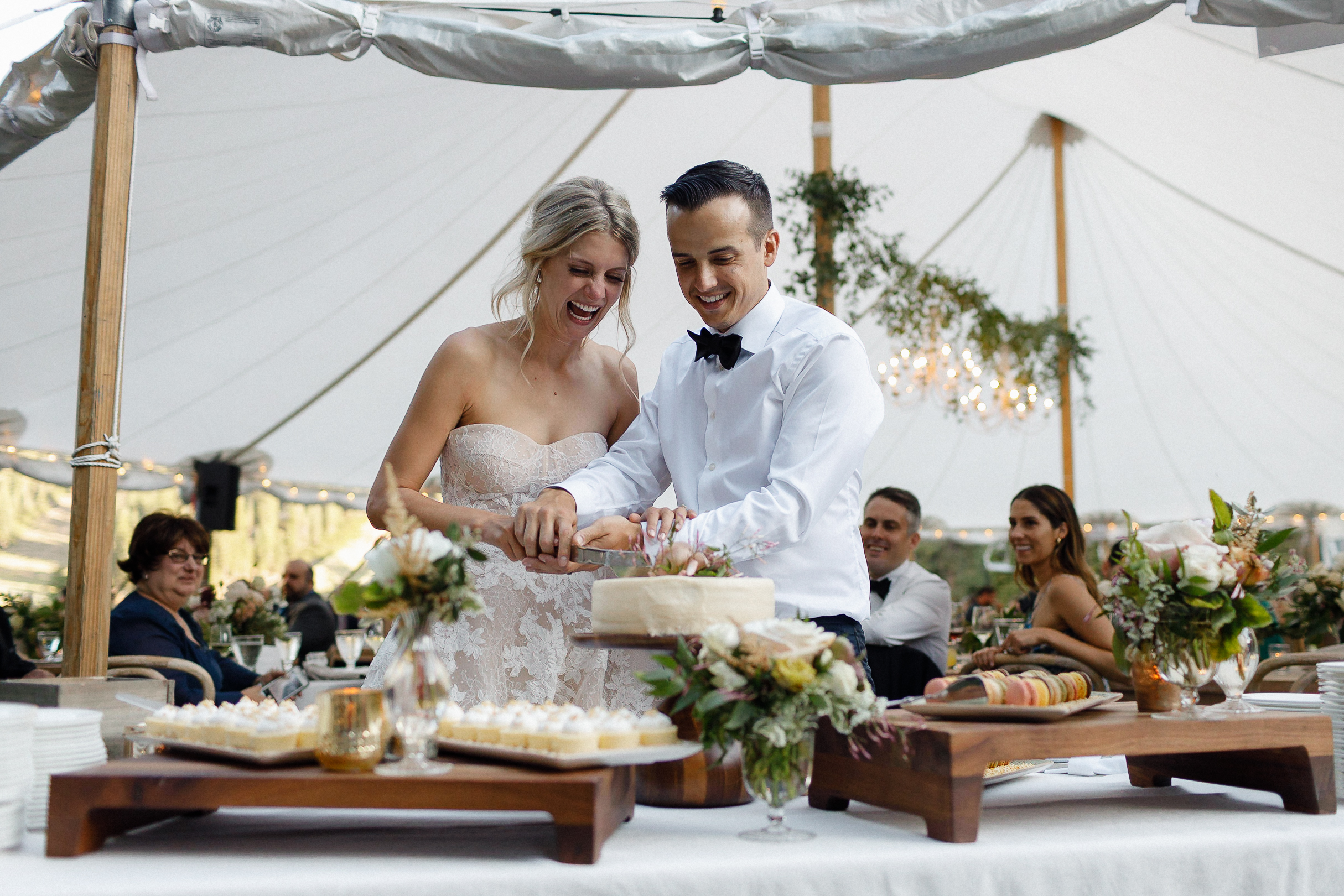 Bride and groom cut their wedding cake together under a white tent at the Ritz-Carlton, surrounded by guests and dessert tables.