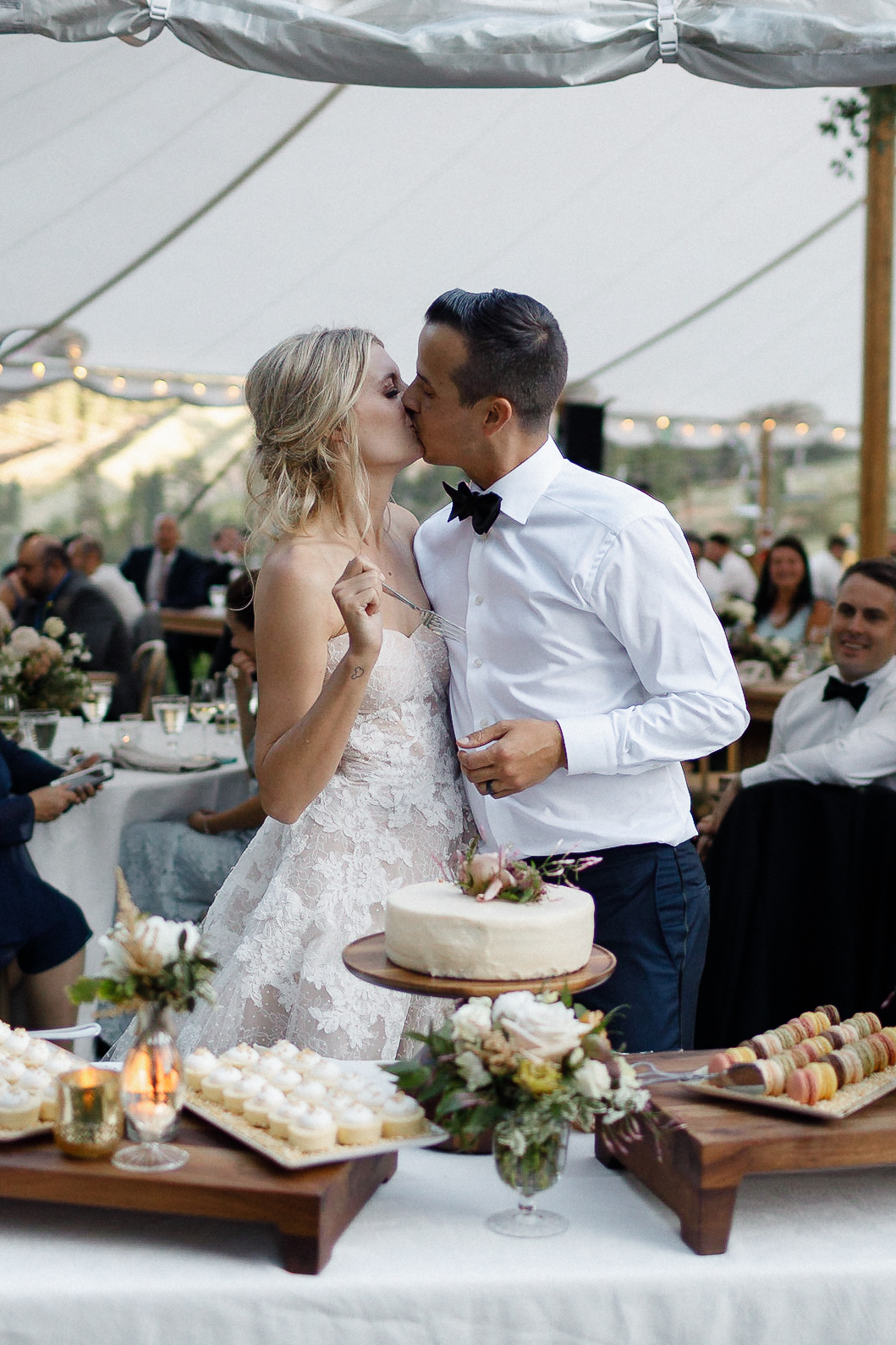 Bride and groom kiss by their wedding cake at a dessert-filled table, with guests celebrating under string lights in a tent.