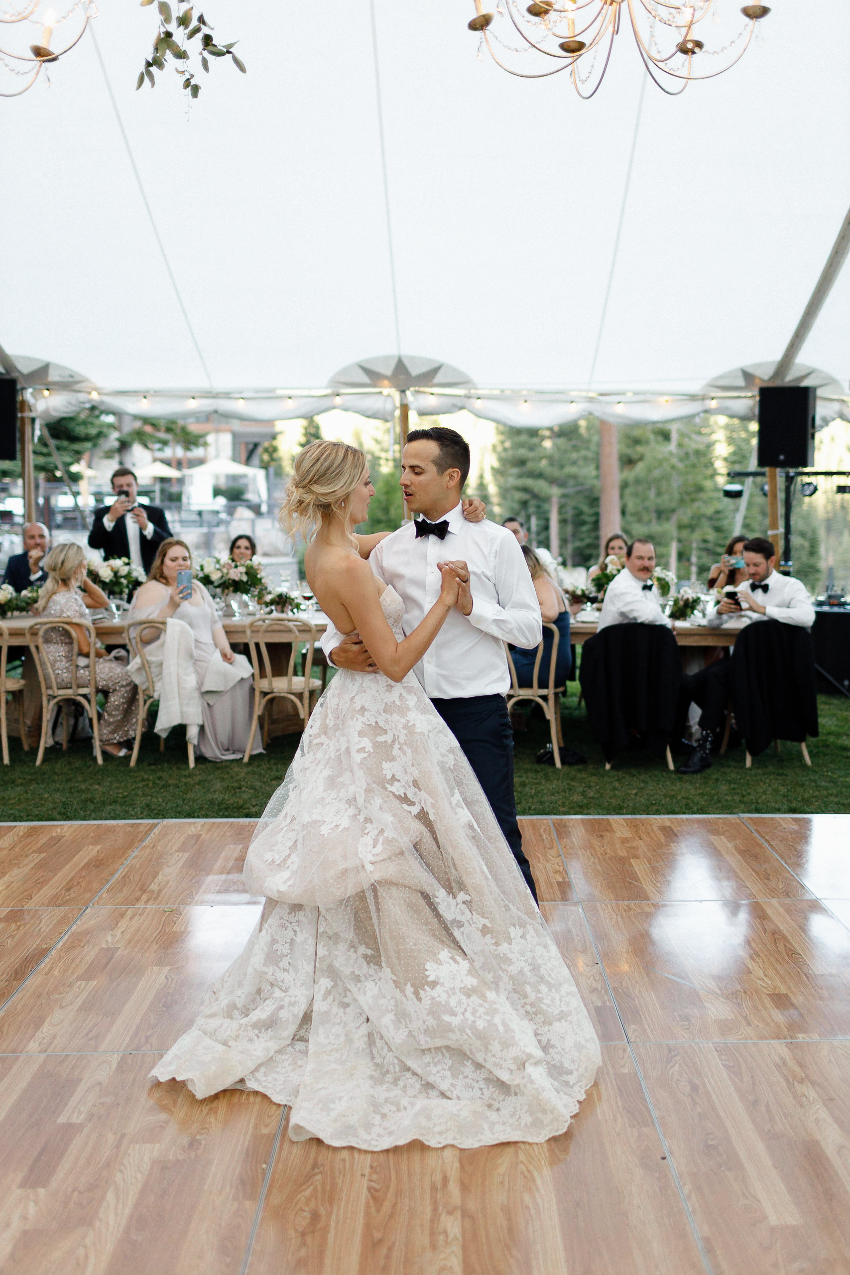 Bride and groom have their first dance on a wooden floor under a white tent, with guests seated at decorated tables outdoors.
