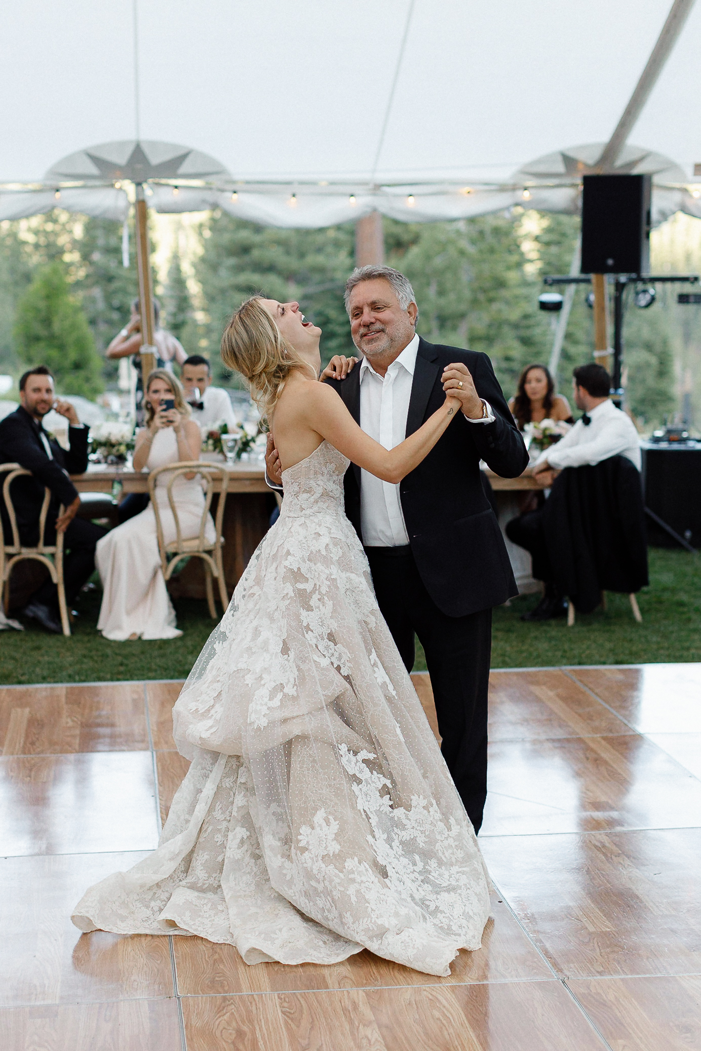 A bride in a white lace gown dances with an older man in a black suit at a Lake Tahoe wedding, guests seated behind them.